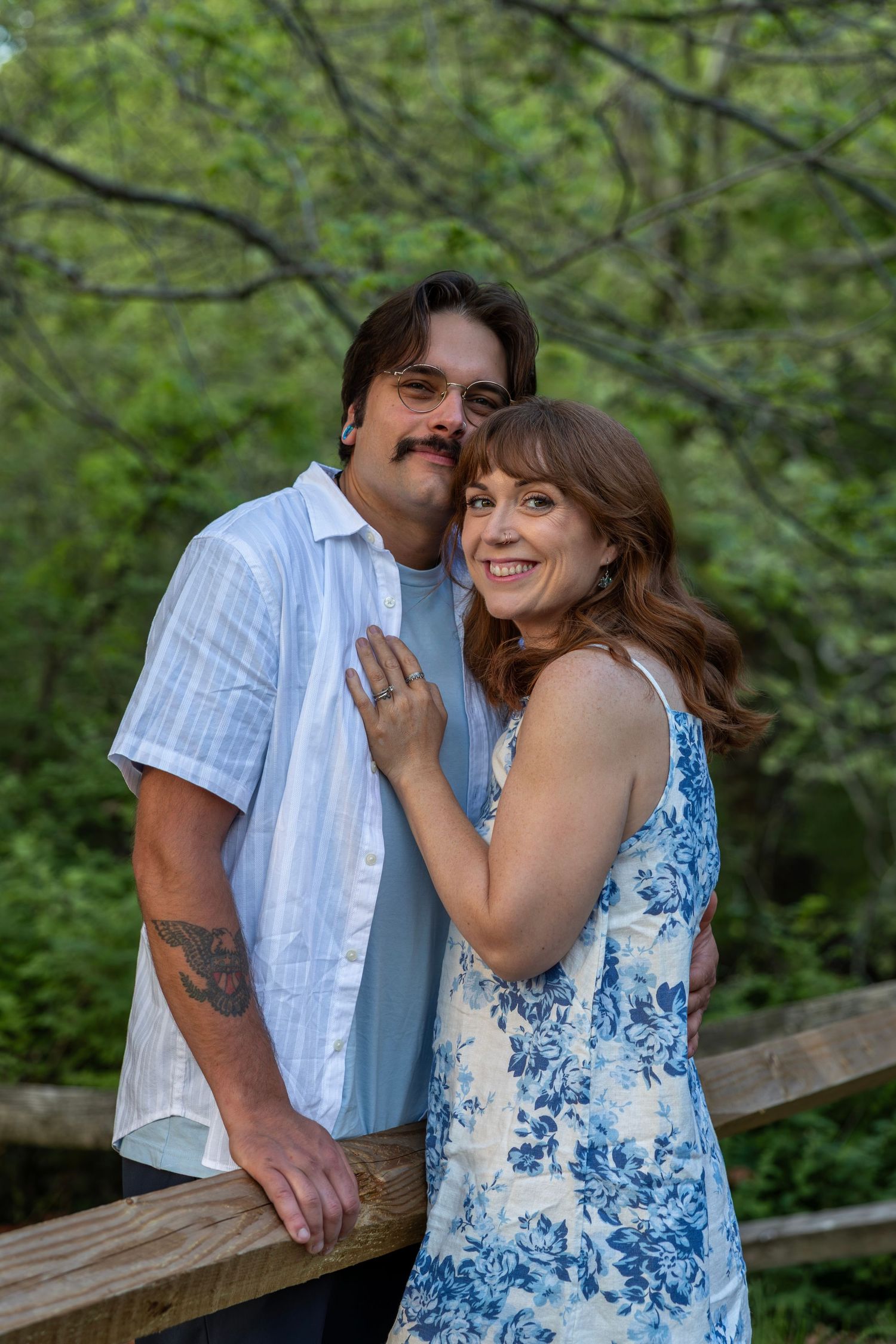Smiling couple poses together on a wooden bridge surrounded by lush green trees in an outdoor portrait.
