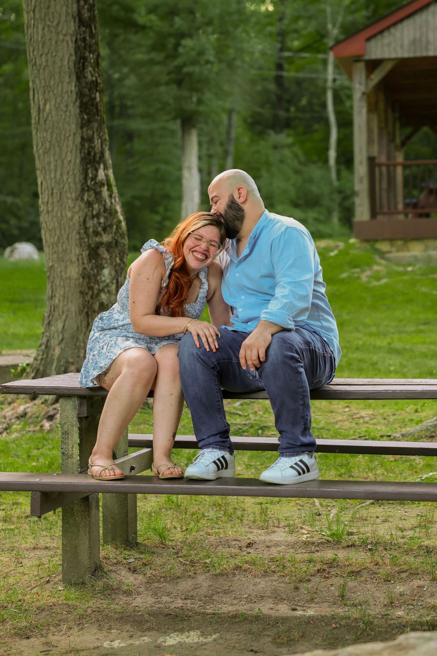 Couple laughing and sharing a whisper while sitting together on a wooden picnic bench in a park.