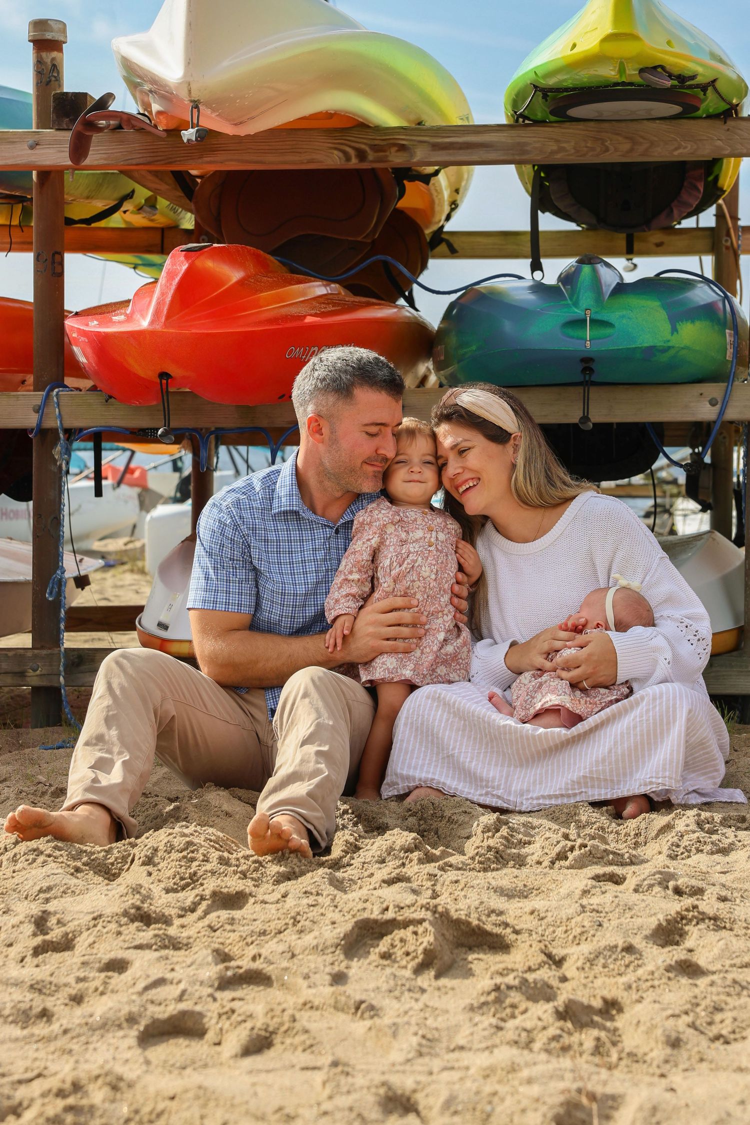 Family of four sitting on sandy beach in front of colorful kayak rack, parents lovingly holding toddler and newborn baby.