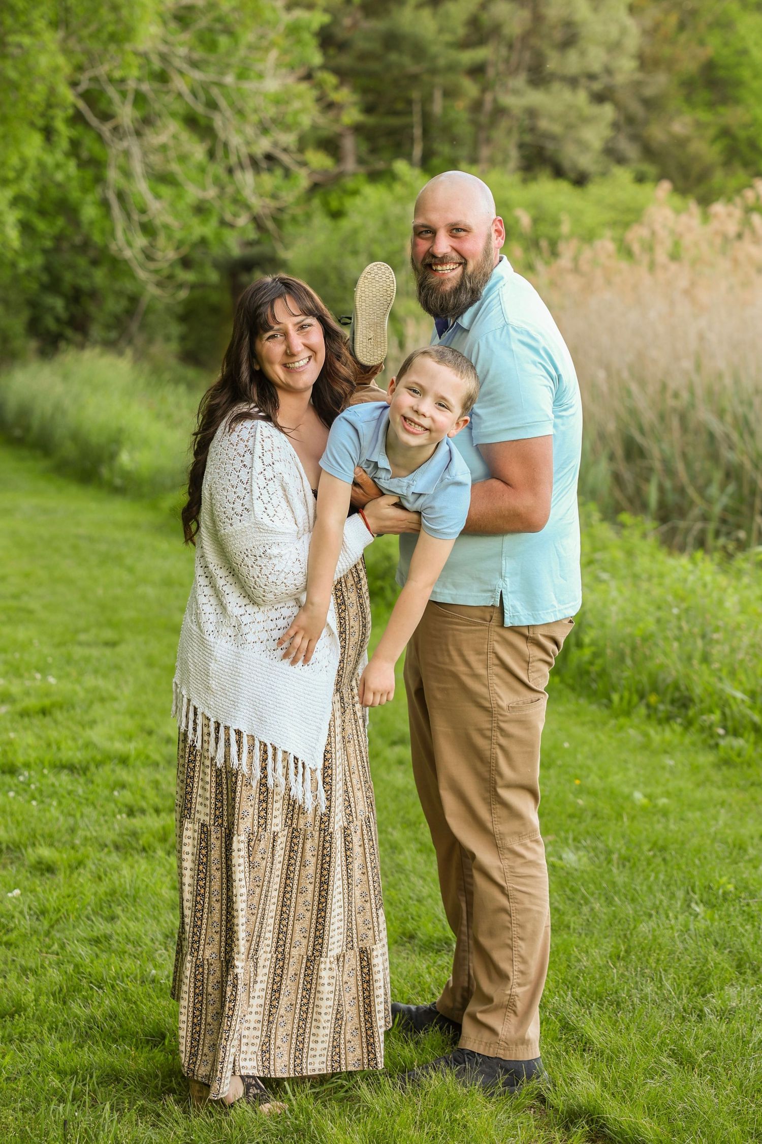 A smiling family of three poses together on a lush green lawn with trees in the background.