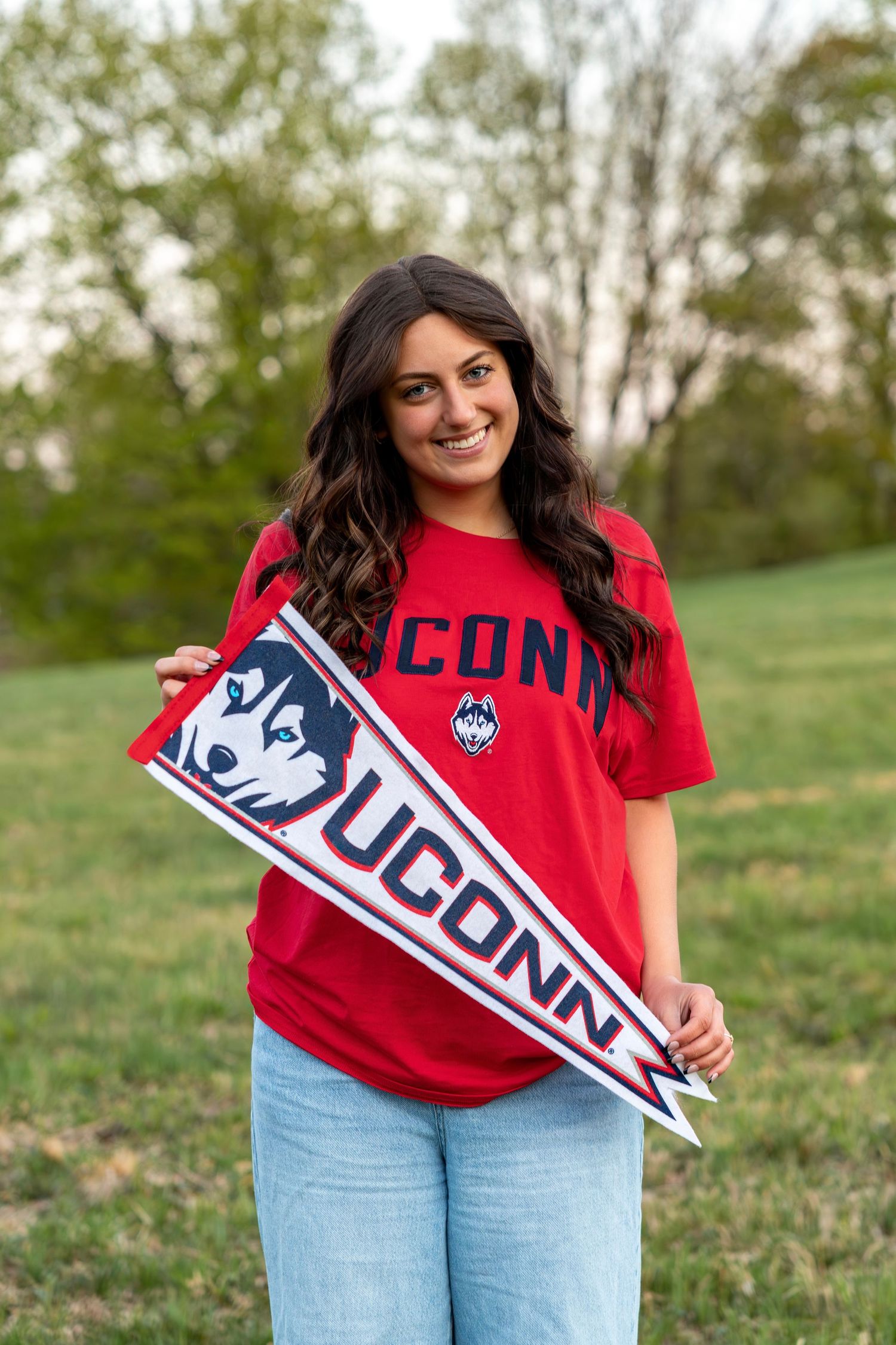 Happy young woman holding UConn Huskies pennant flag wearing matching red UConn t-shirt outside.