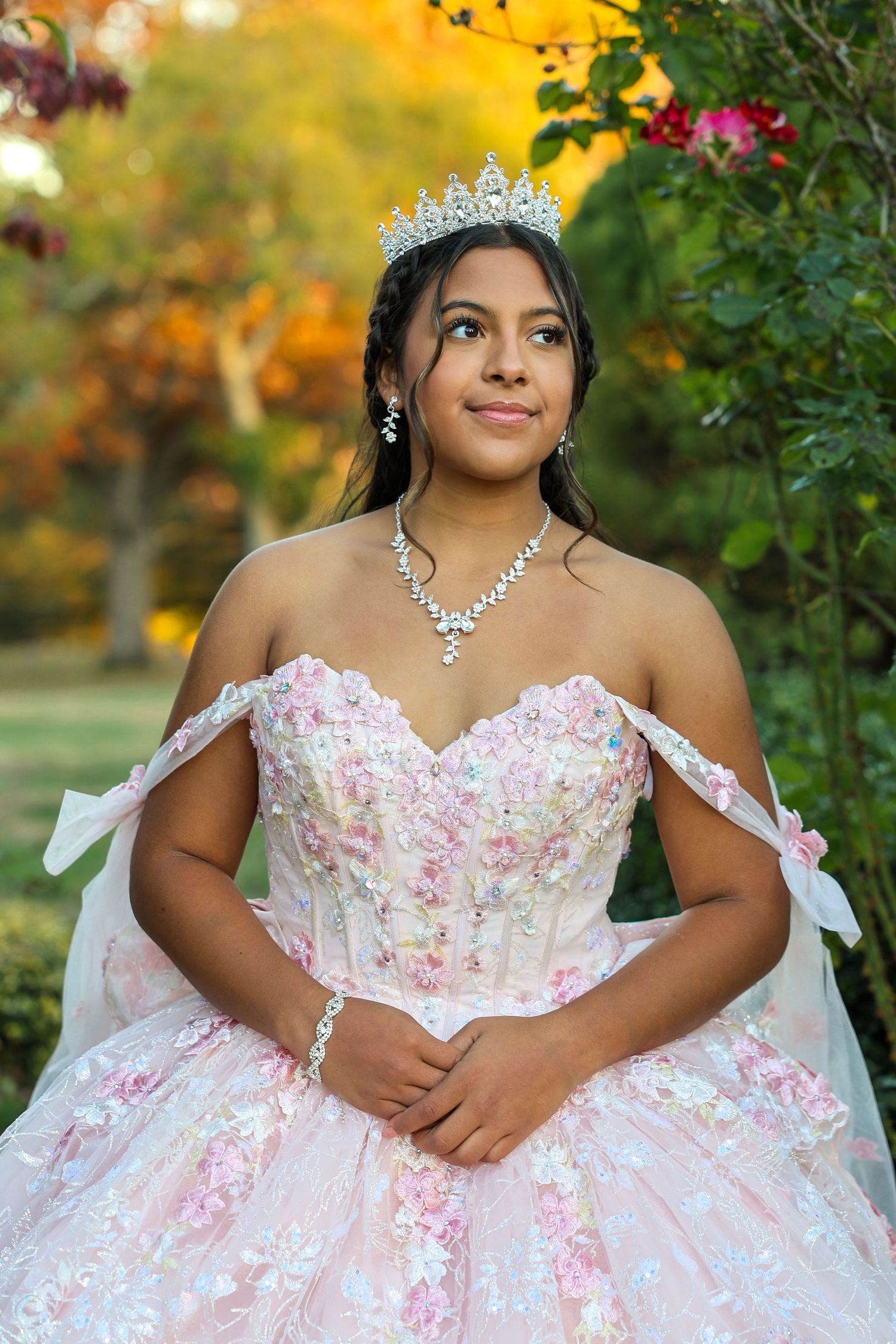Teen girl in pink off-shoulder floral quinceañera gown with crystal tiara gazing upward in garden.