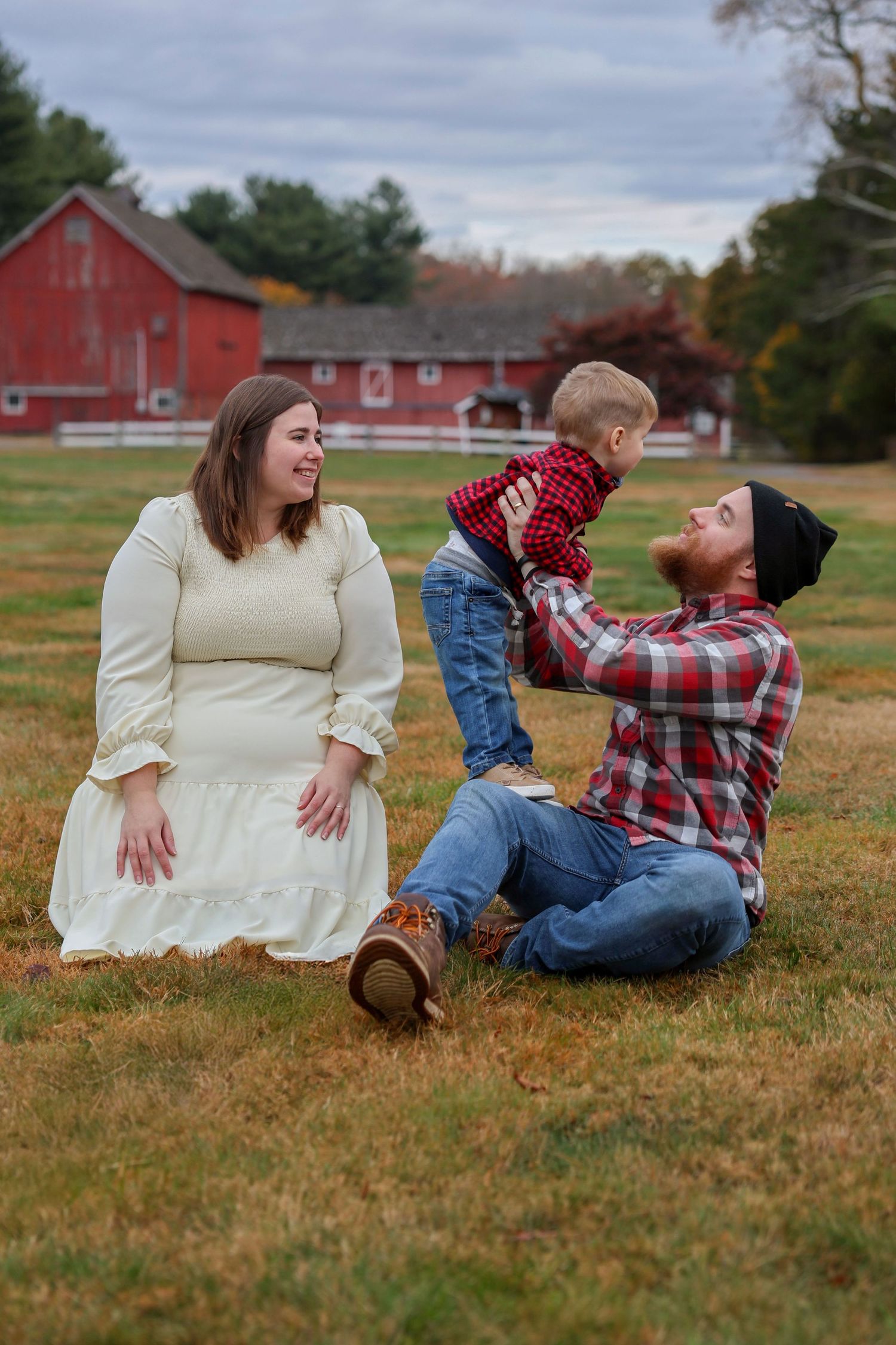 Family sitting on autumn grass near red barn, father lifting laughing toddler while mother watches warmly.