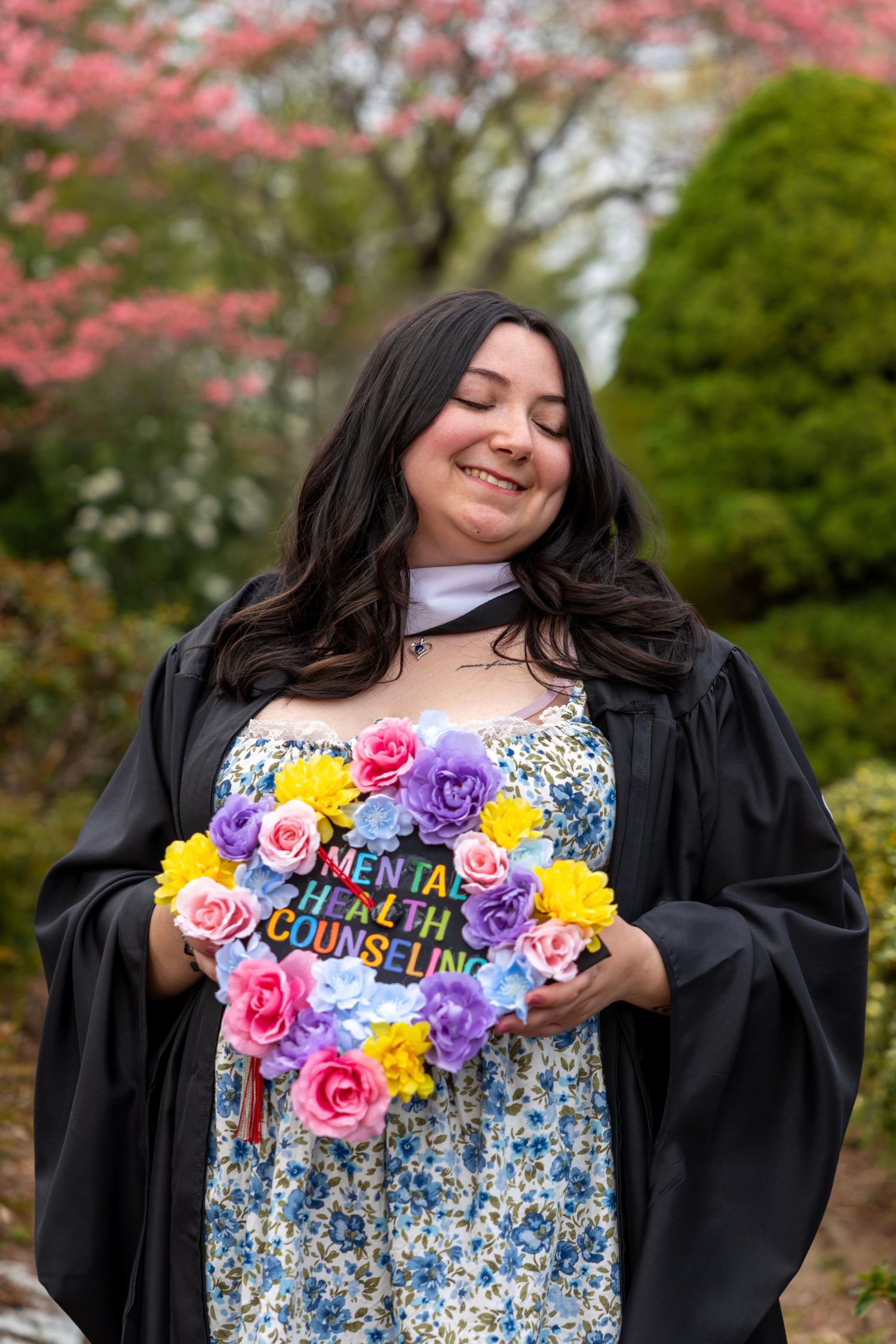 Graduate in black gown holds decorated cap with colorful flowers reading Mental Health Counseling in spring garden.