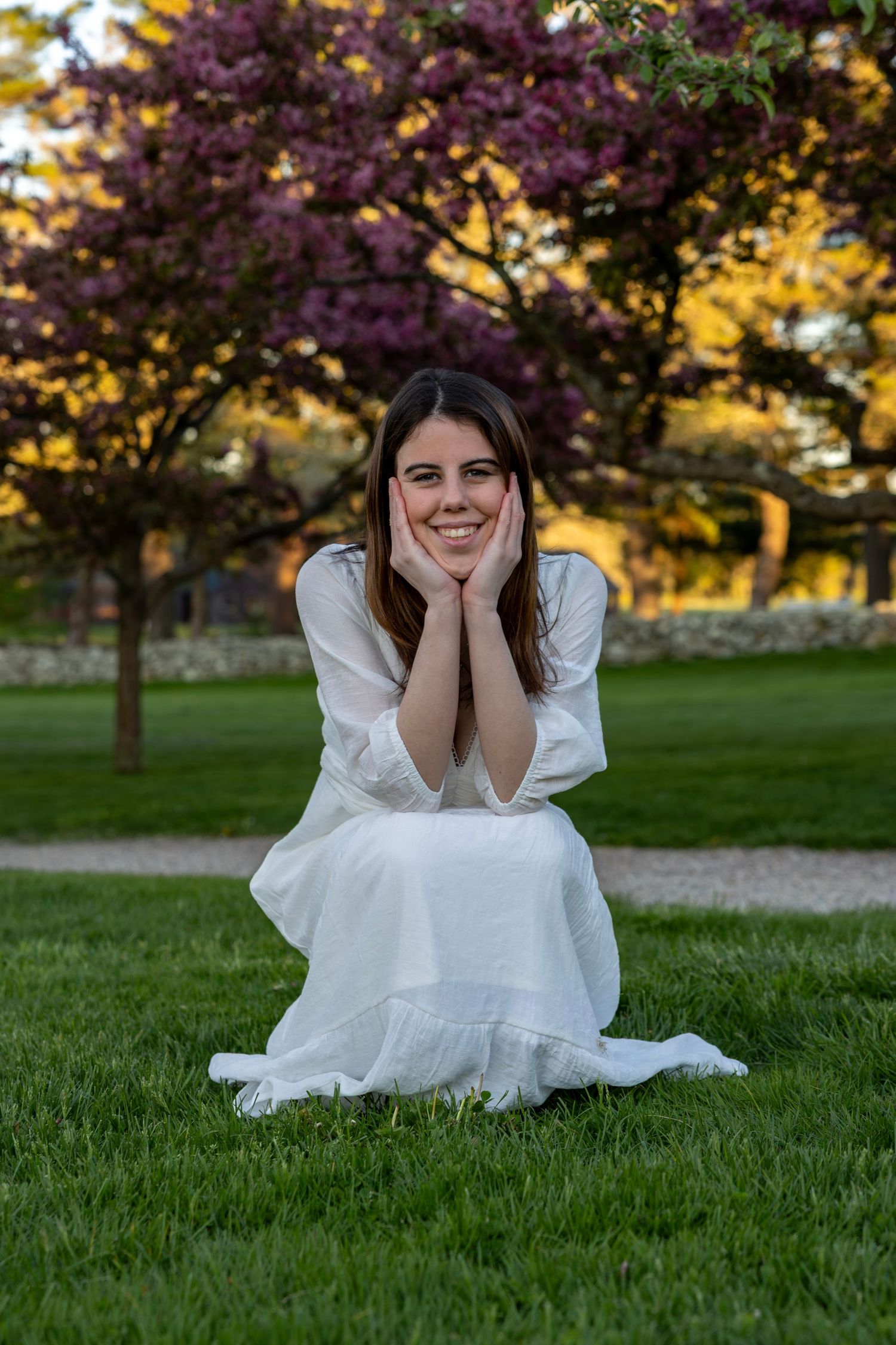 Joyful woman in white dress kneeling on grass with hands on cheeks near trees.