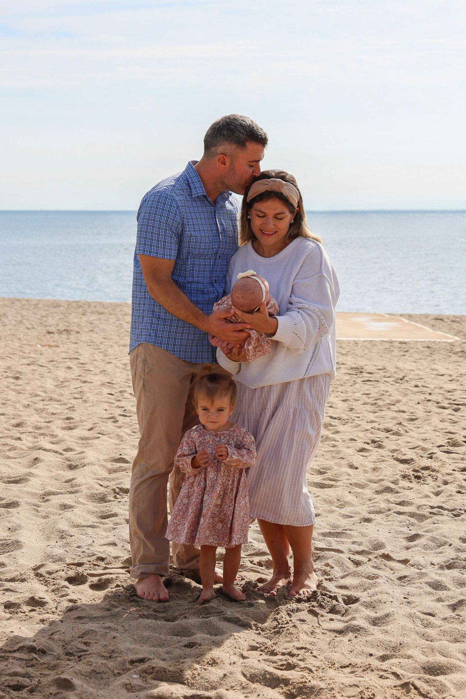 Happy family with father, mother, newborn baby, and toddler daughter pose together on sandy beach.