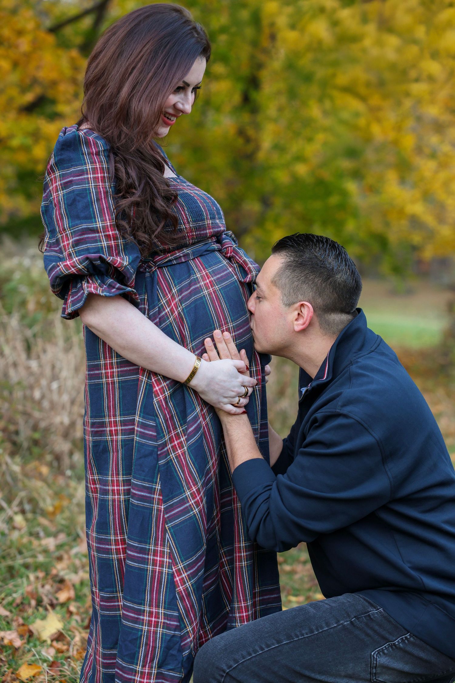 Man kneeling to kiss pregnant womans baby bump in autumn park with golden yellow foliage background.