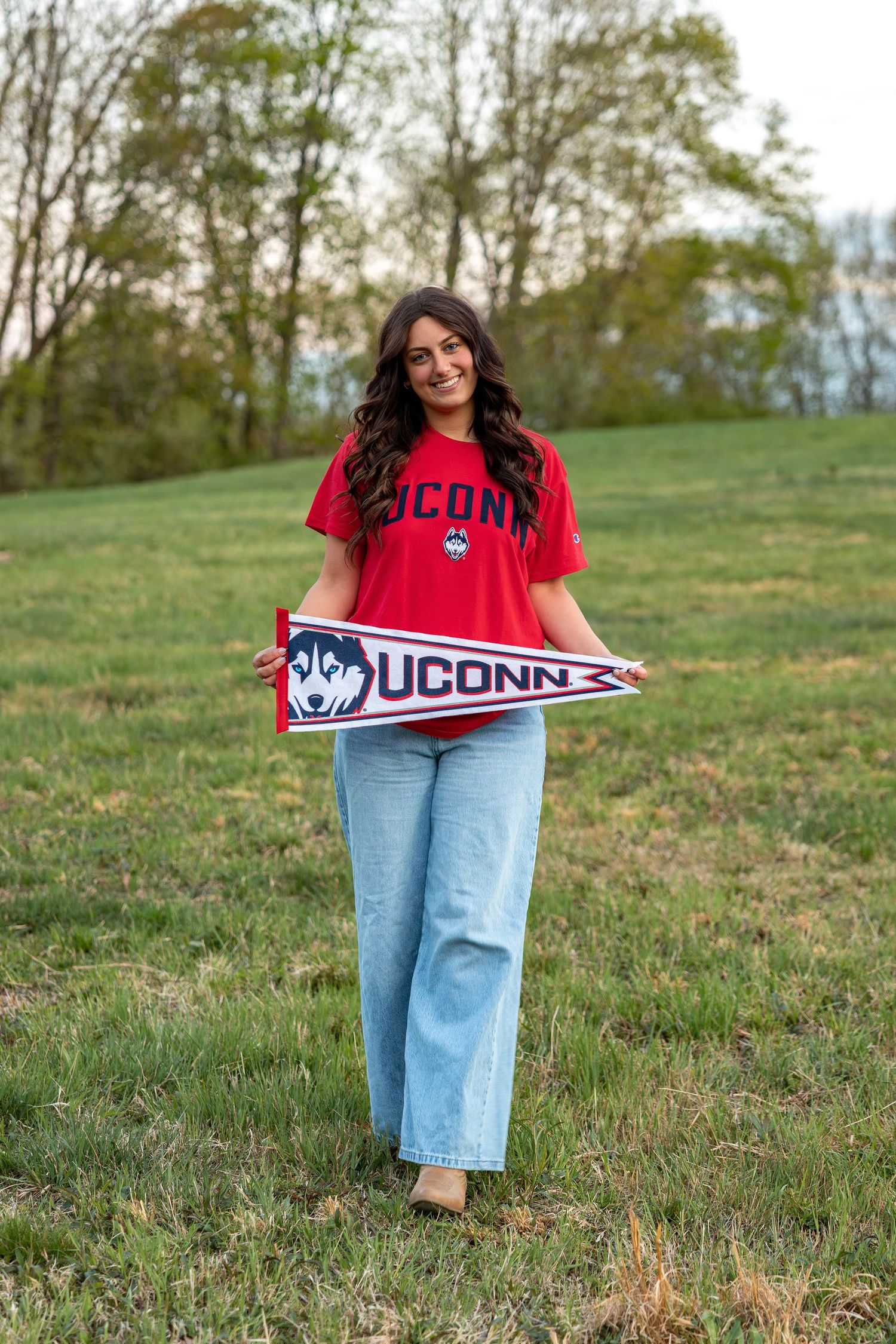 Smiling woman walking in green field holding UConn Huskies pennant in red t-shirt and light jeans.