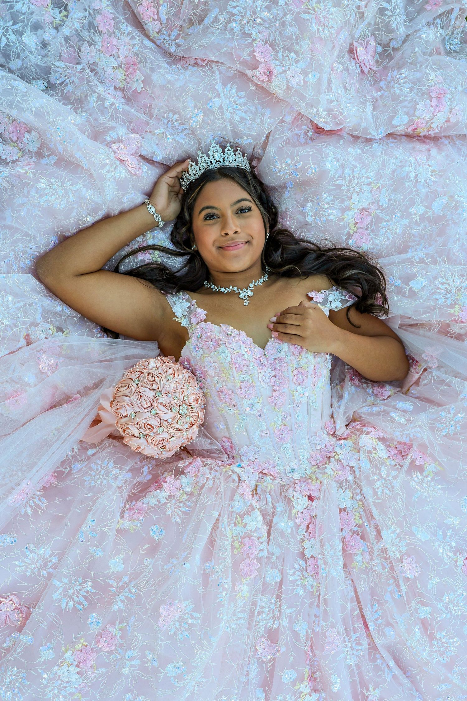 Teen girl in pink floral quinceañera gown lying down holding a pink rose bouquet with tiara.