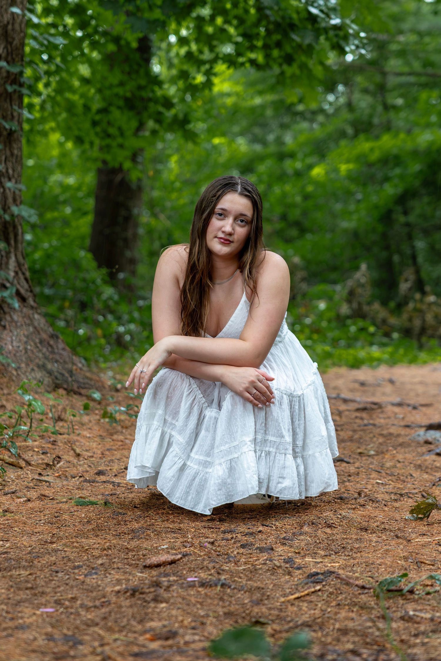 Young woman in white sundress crouching on forest path surrounded by lush green trees.