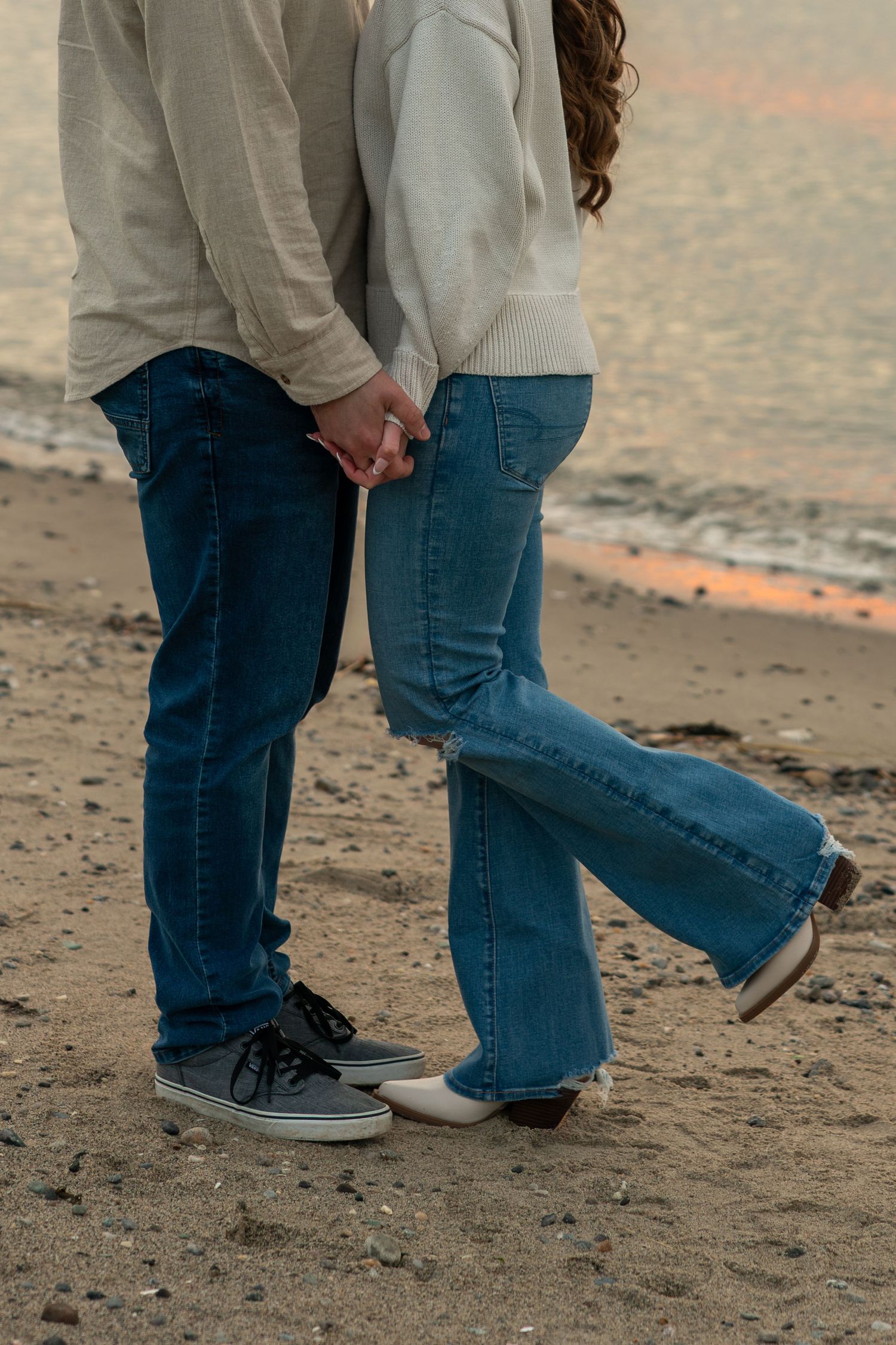 Close-up of couple holding hands in jeans and sneakers on sandy beach at sunset.