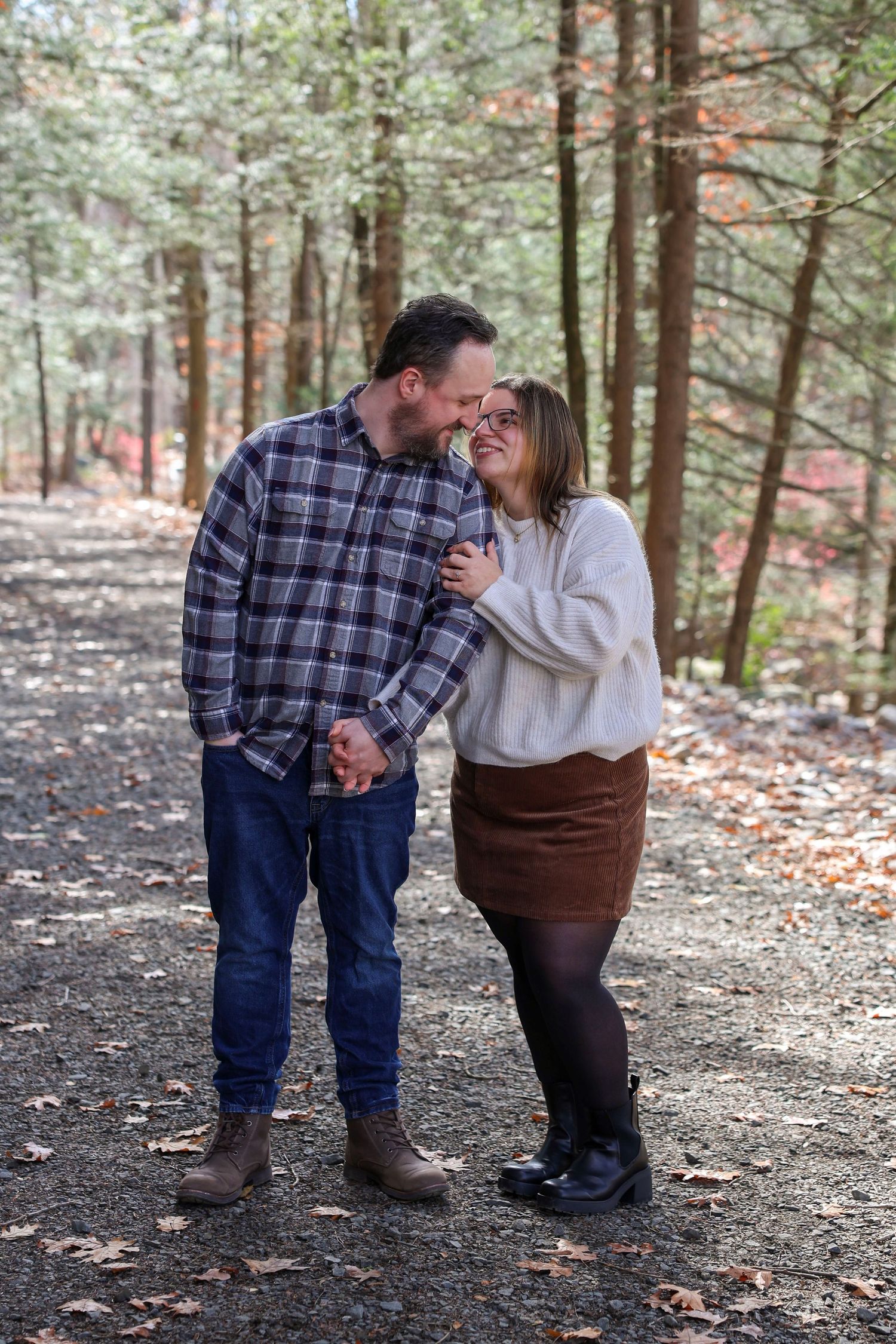 A couple shares a romantic moment on a leaf-covered forest path, smiling and gazing at each other.