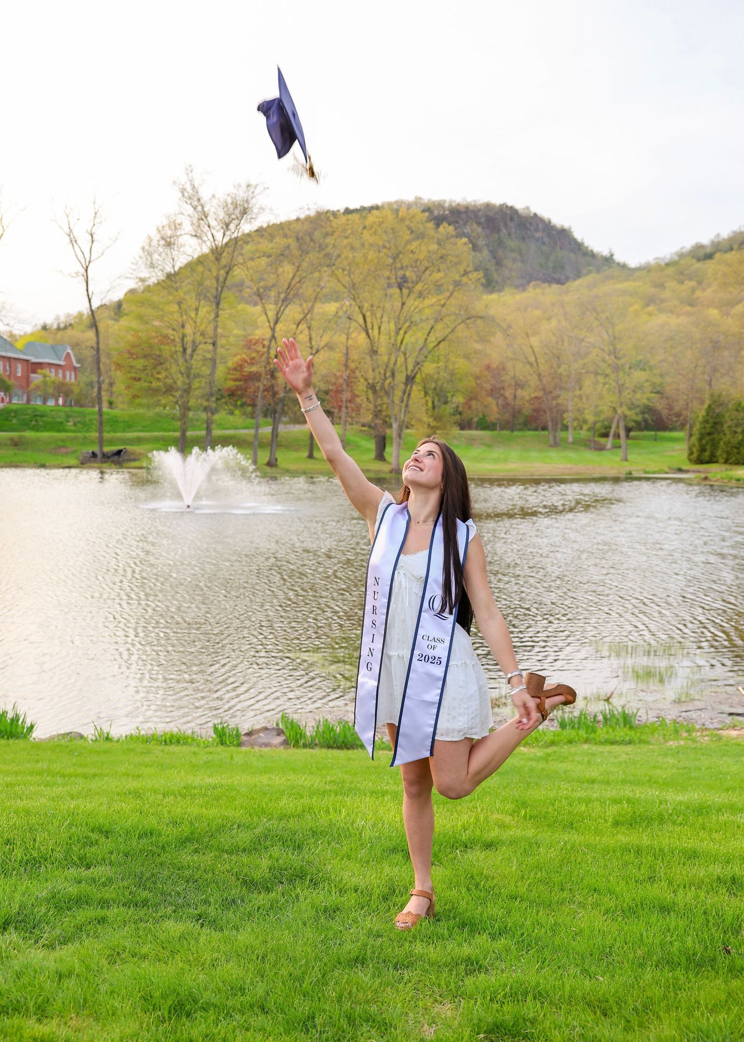 Nursing graduate tossing cap in air by scenic pond with fountain and mountain backdrop.