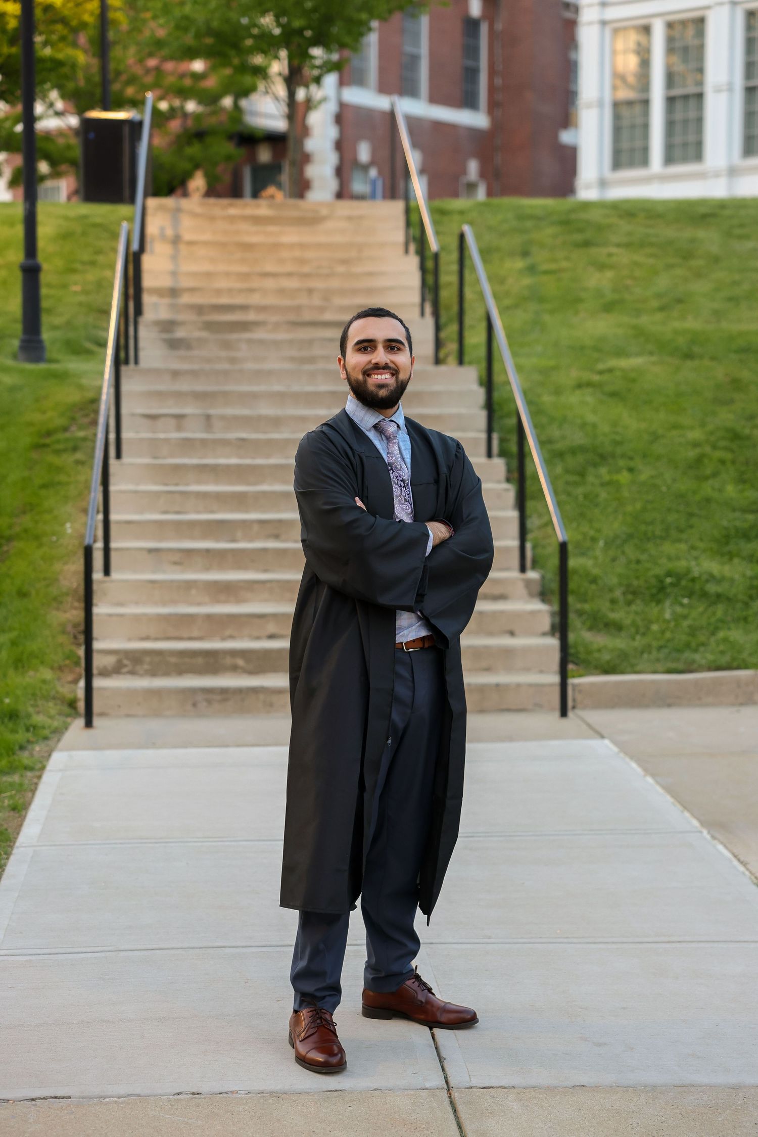 Male graduate in black academic gown standing with arms crossed on campus steps.