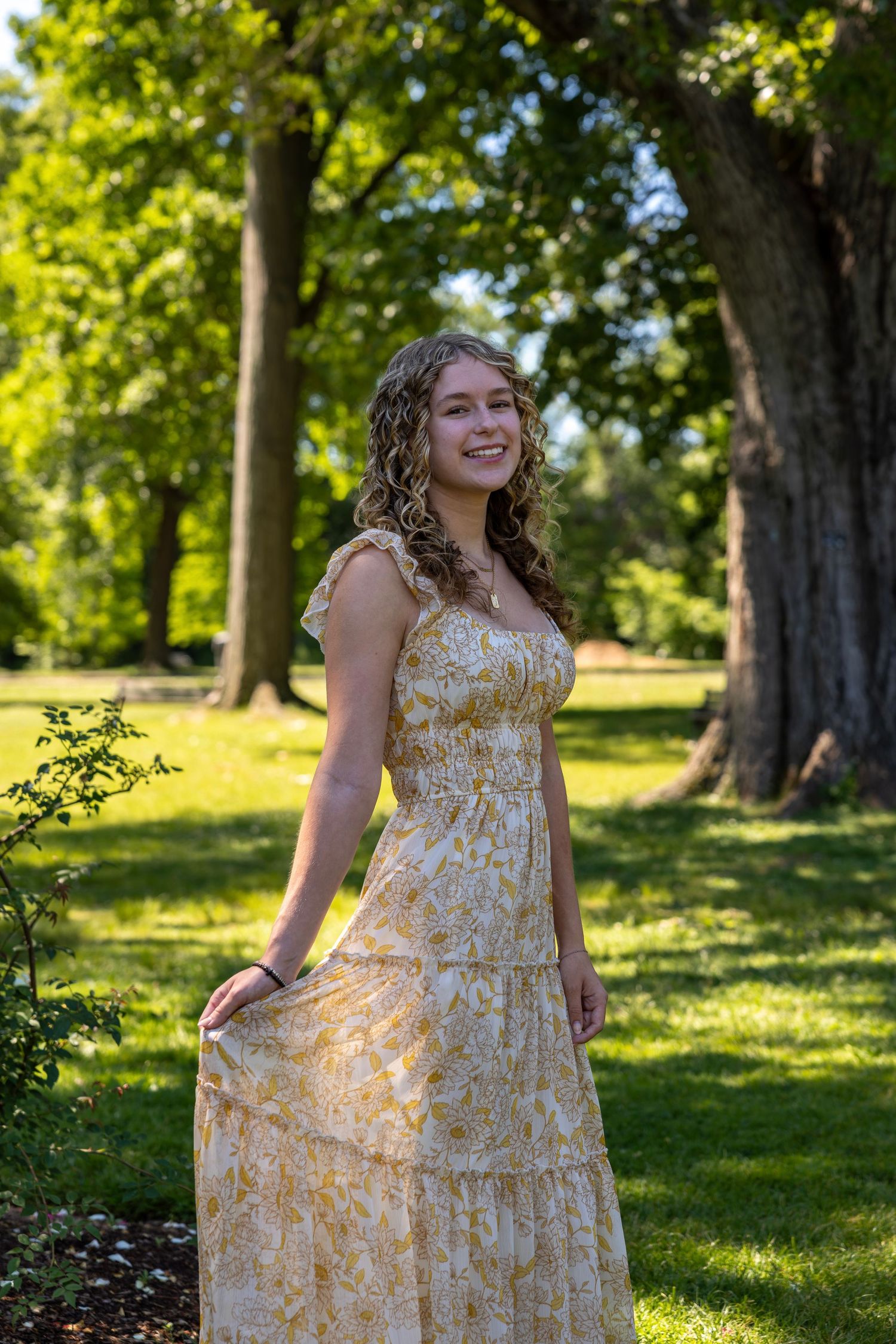 Smiling young woman in yellow floral dress holding skirt while posing in a lush green park setting.