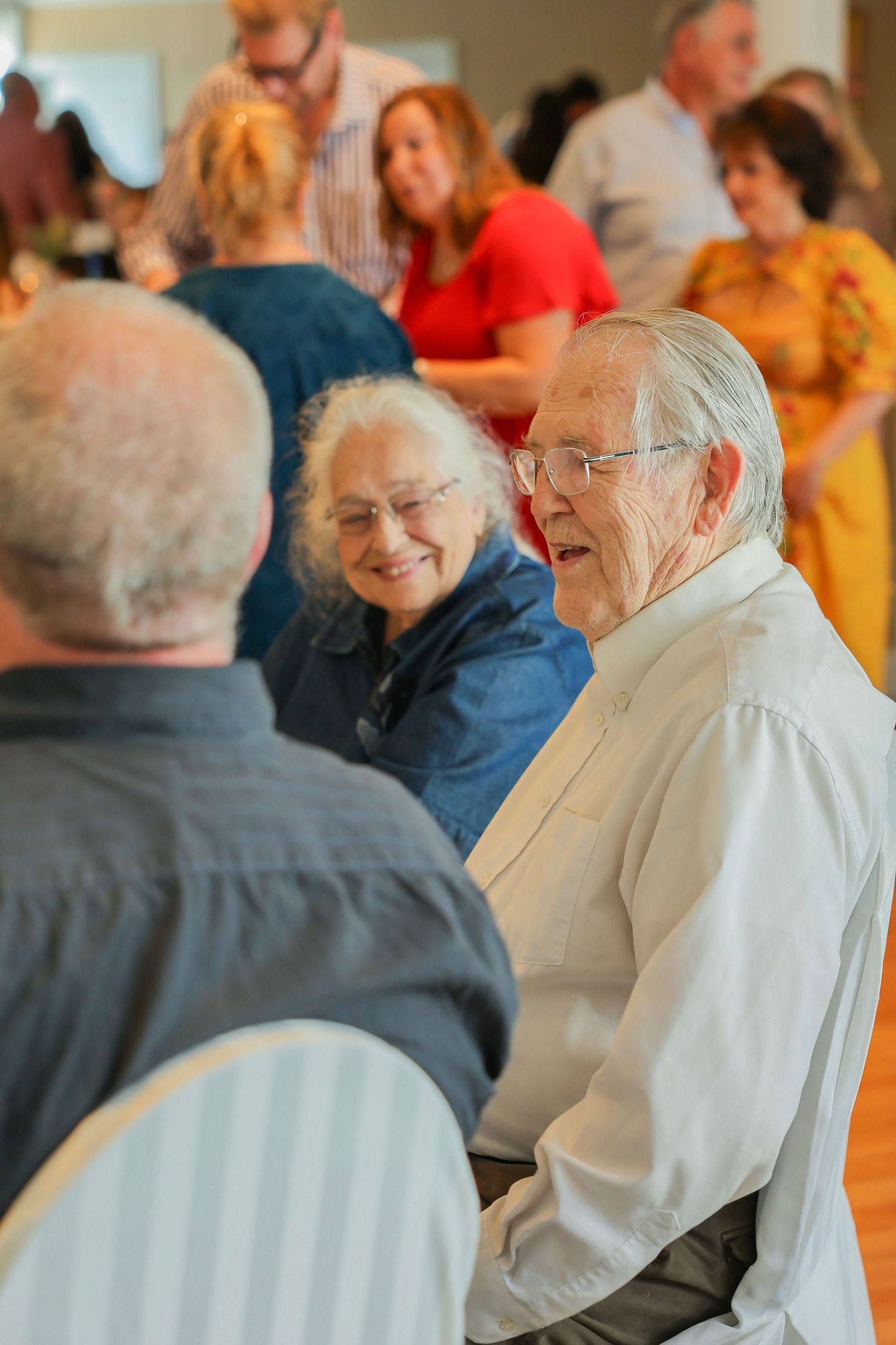 Elderly couple smiling and conversing with guests at a lively indoor celebration event.