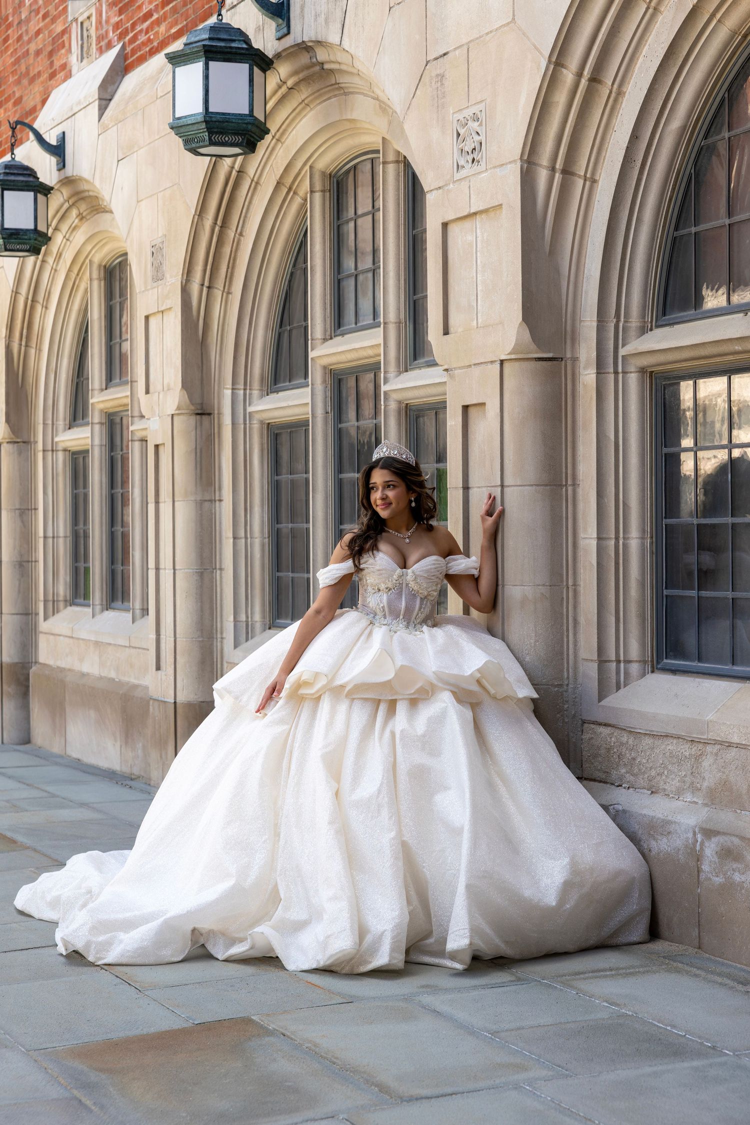 Quinceañera in white tiered ball gown with crown posing under stone arched colonnade.