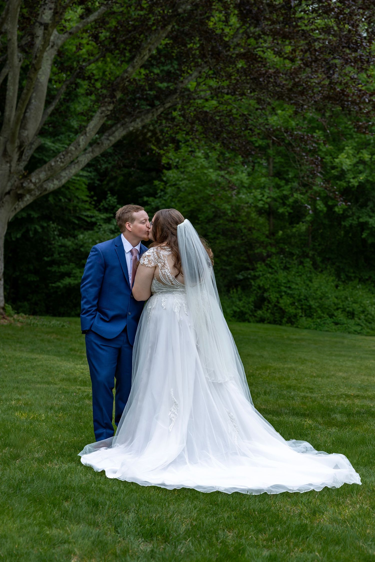 Bride and groom share a romantic kiss on lush green lawn with trees in background on their wedding day.