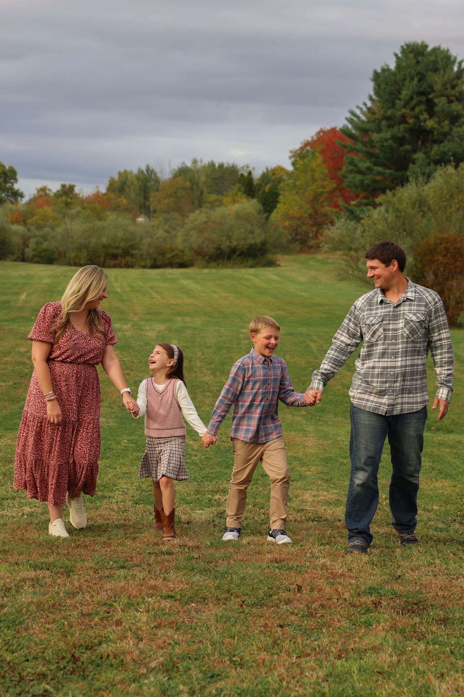 Happy family of four holds hands walking through green field with fall foliage background.