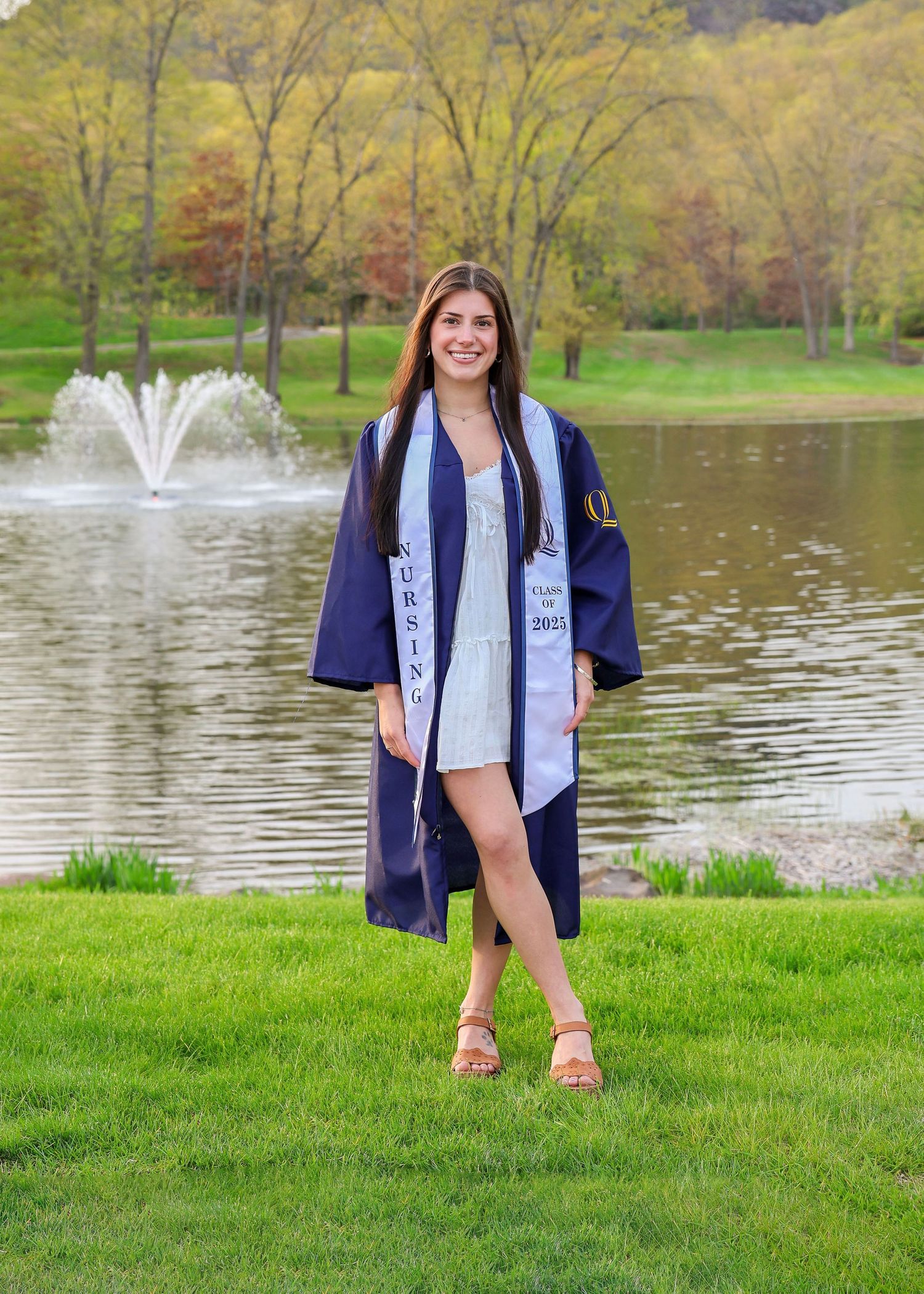 Smiling nursing graduate in navy cap and gown poses by a scenic pond with fountain on a sunny spring day.