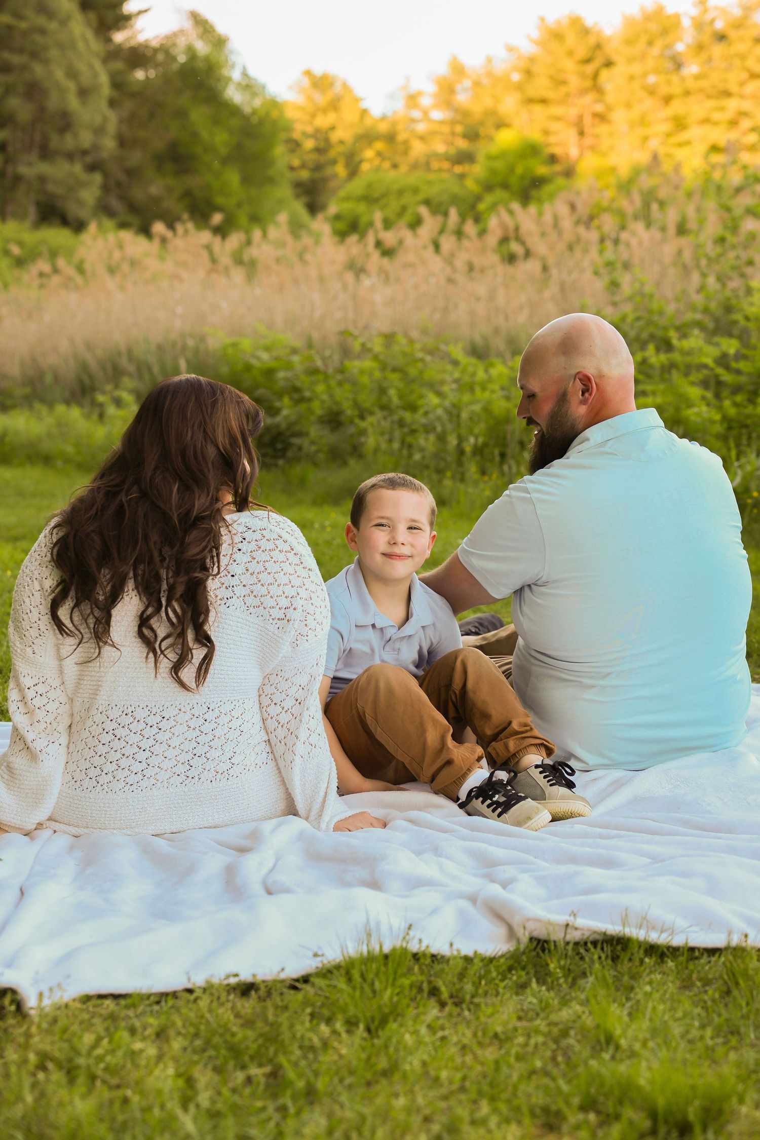 A happy family of three sits on a white blanket in a sunny green meadow near tall grass.