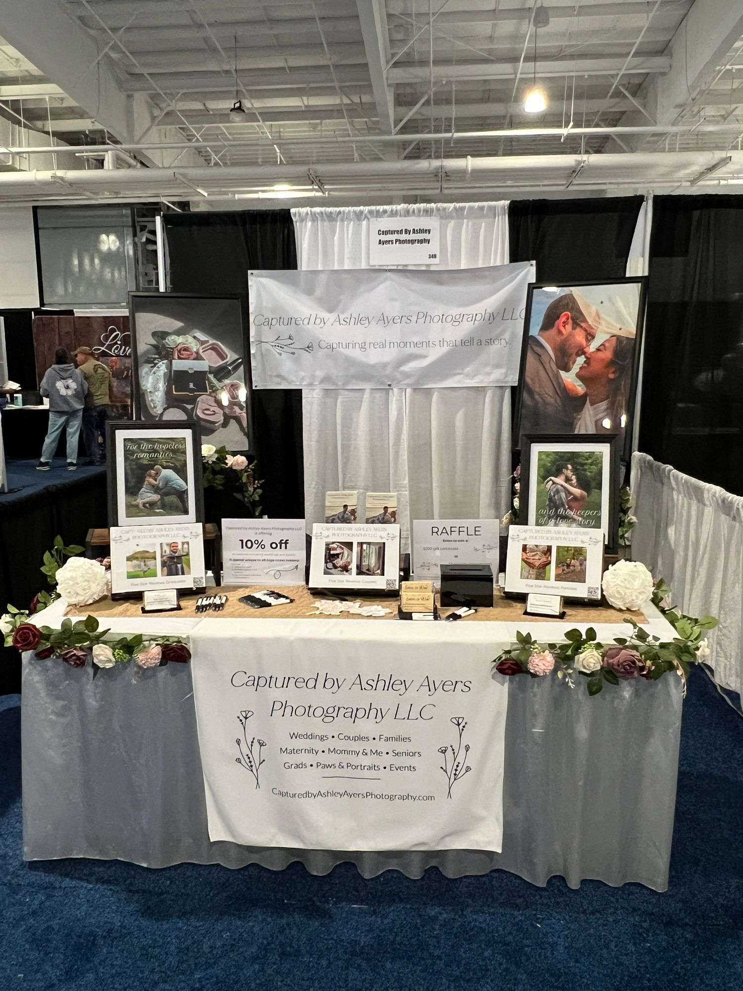 A professional photography vendor booth for Captured by Ashley Ayers Photography LLC decorated with floral arrangements and framed prints.