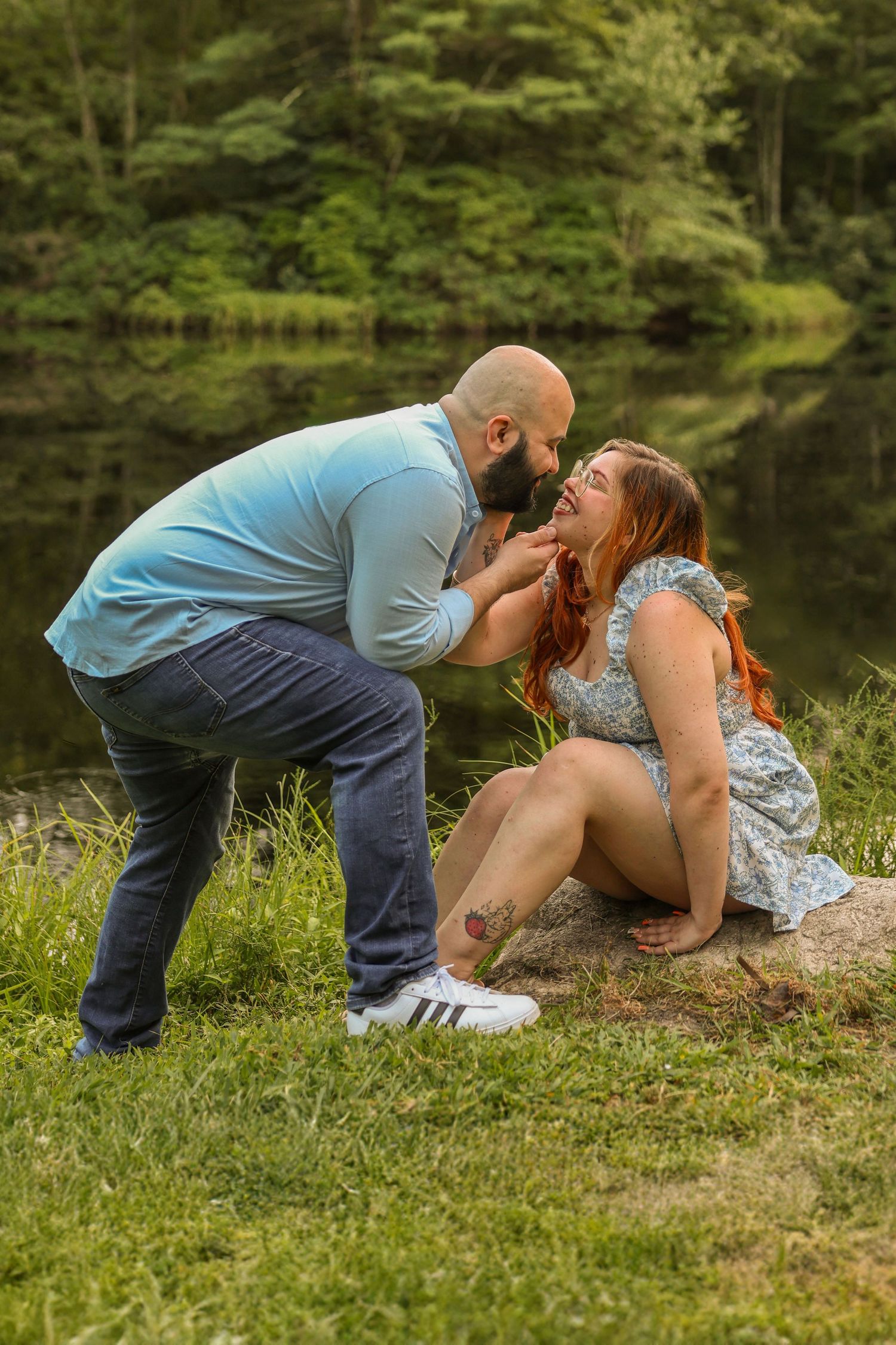 Man feeding his laughing redhead girlfriend a strawberry while sitting on a rock by a forest lake.