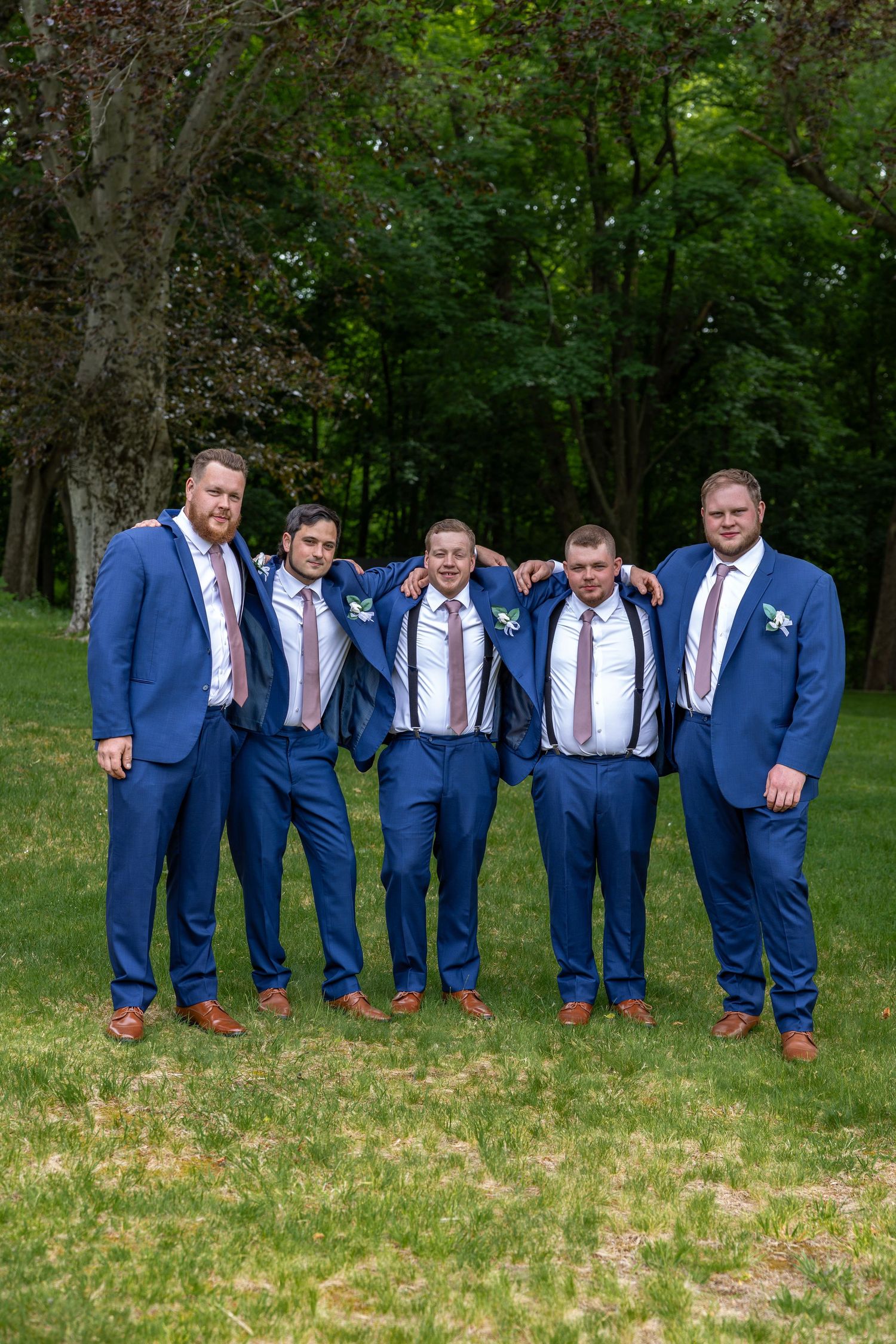 Five groomsmen in navy blue suits with pink ties pose together on green grass at wedding.