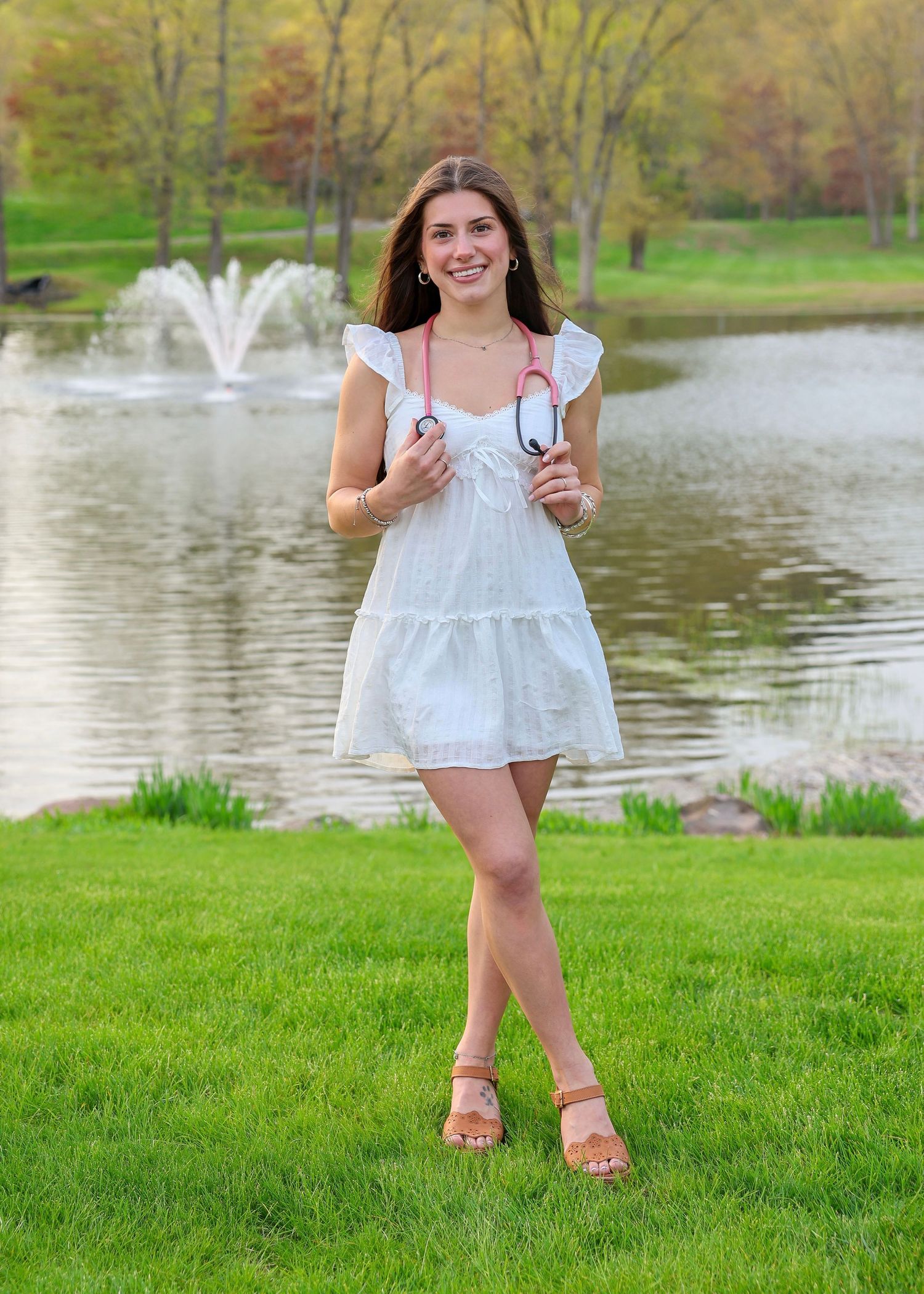 Smiling nursing graduate in white dress holding stethoscope while walking on green lawn by pond.