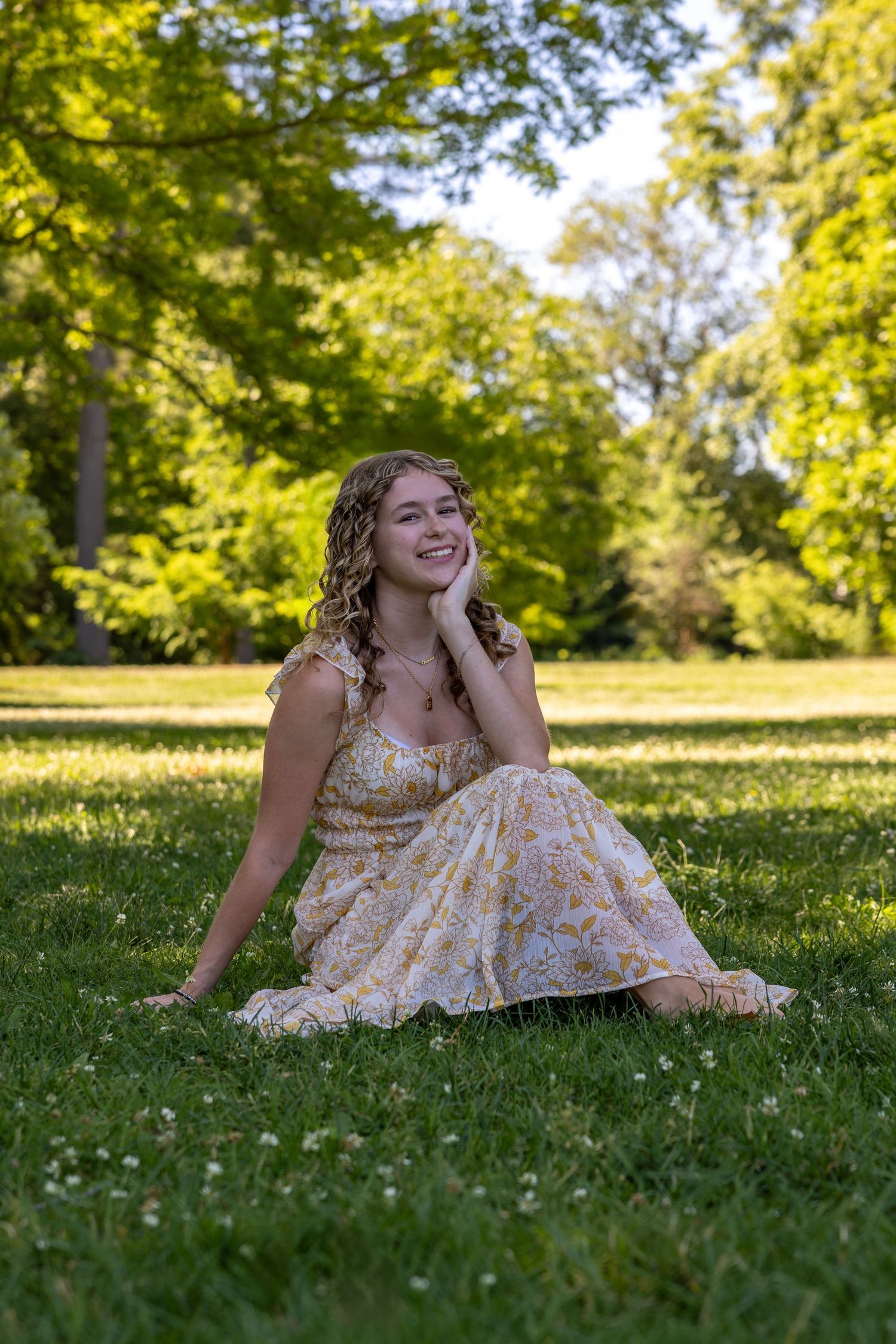 Young woman in yellow floral maxi dress sitting on grass in a sunny park, smiling and resting chin on hand.