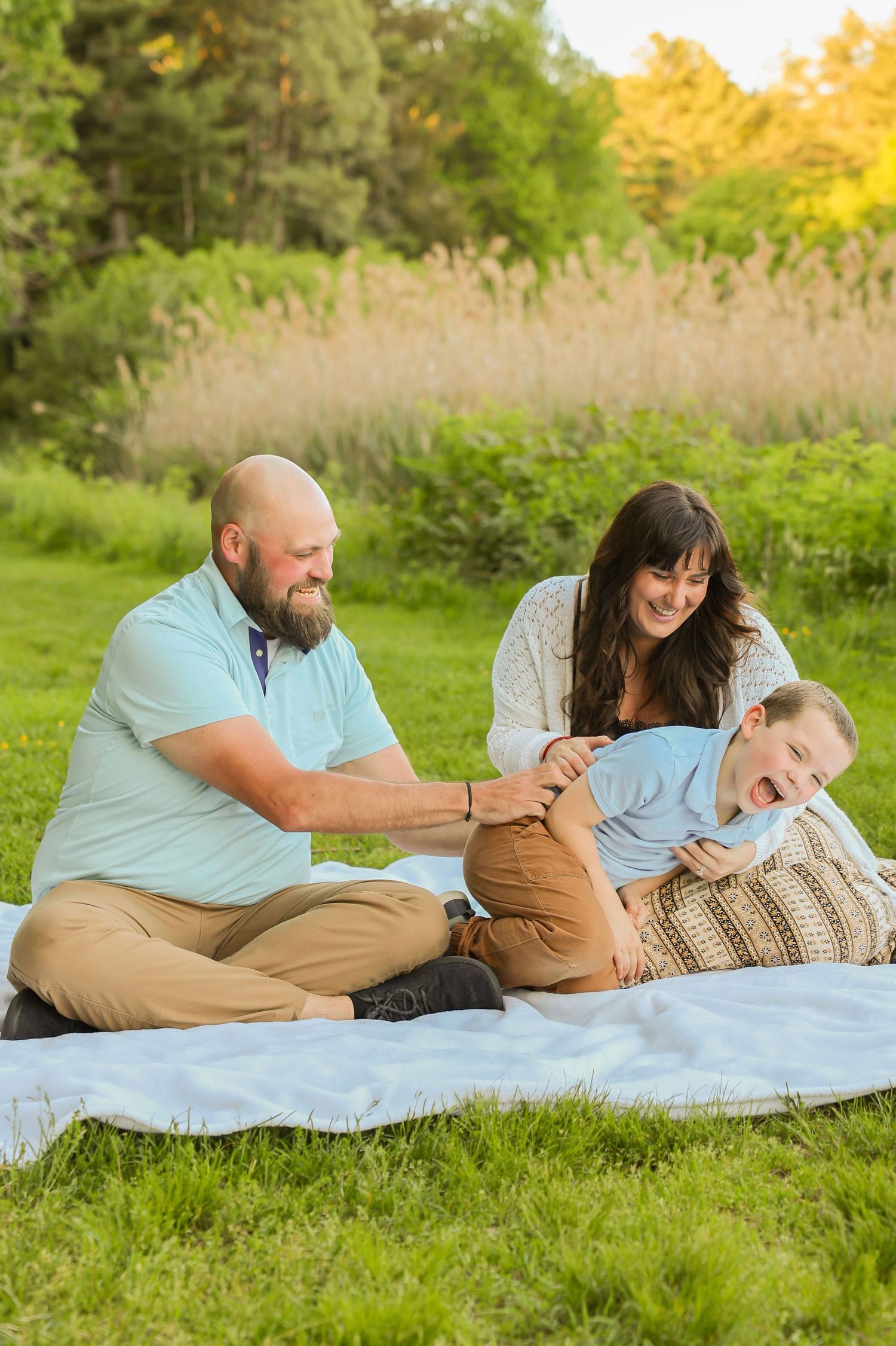 Joyful family of three playing and laughing together on a blanket in a sunny green outdoor park.