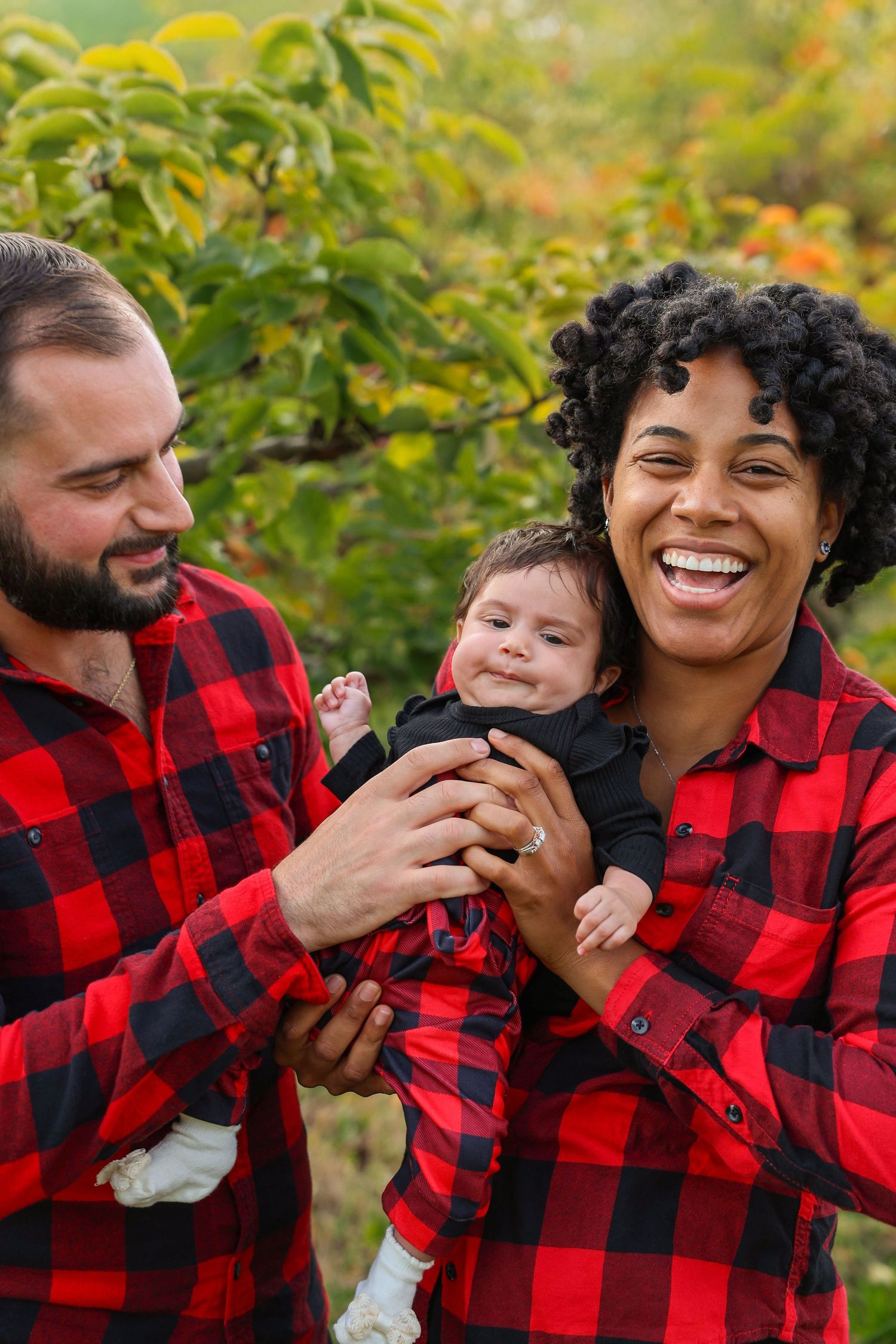 Laughing interracial couple in red plaid shirts holding smiling baby outdoors surrounded by green and yellow foliage.