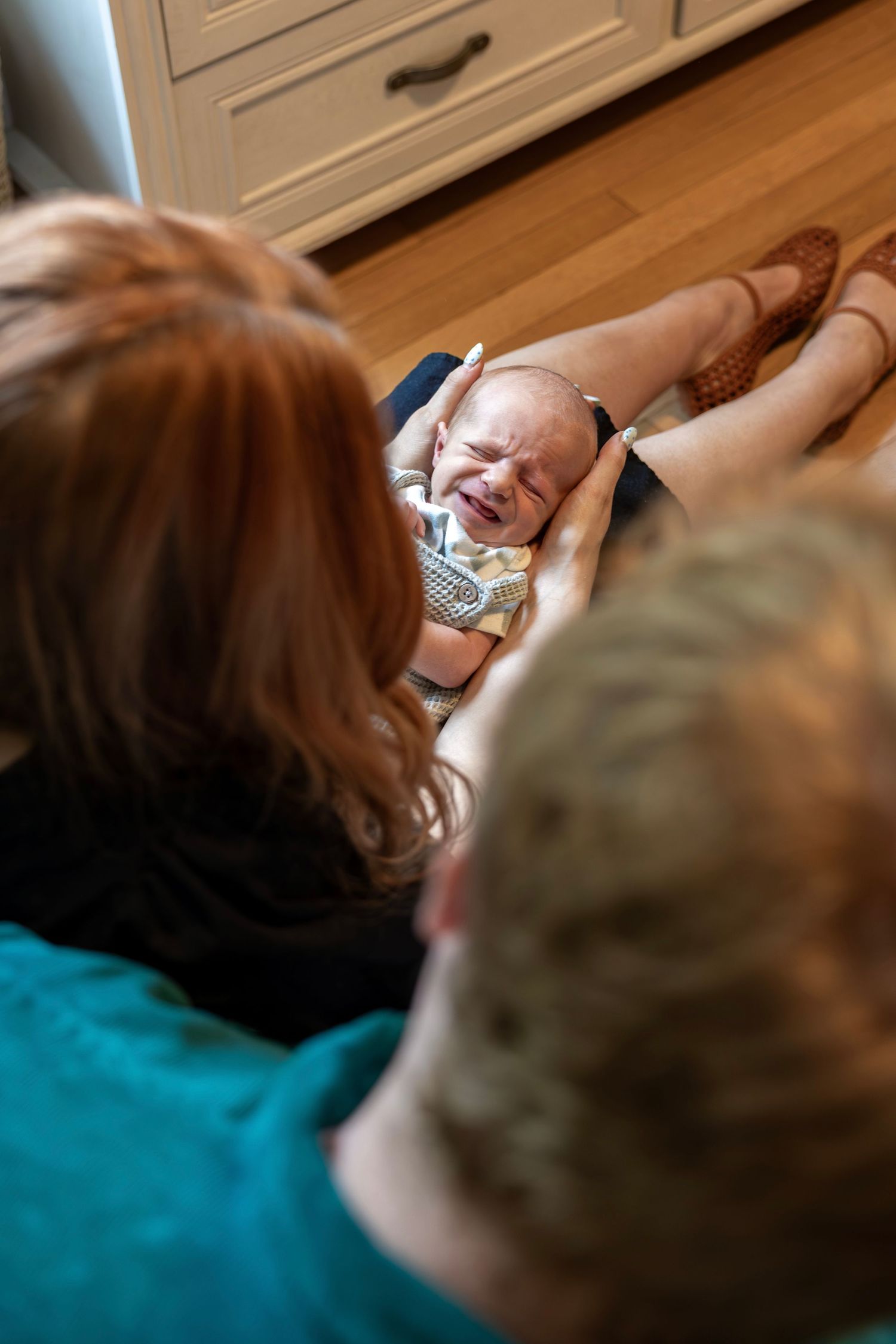 Two parents looking down at their crying newborn baby held in their laps on a wooden floor.