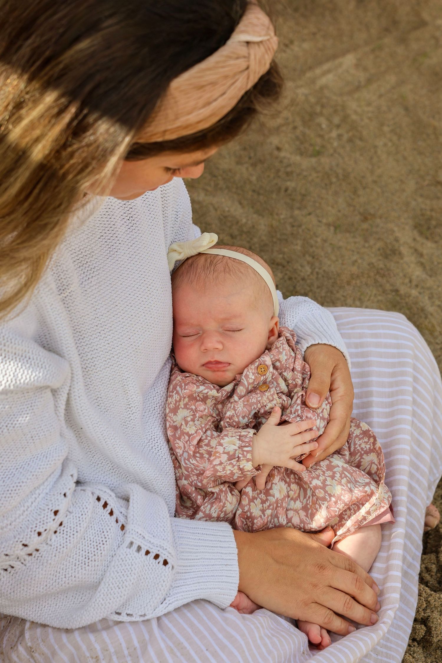 Mother in white knit sweater tenderly holds sleeping newborn baby in floral outfit outdoors.