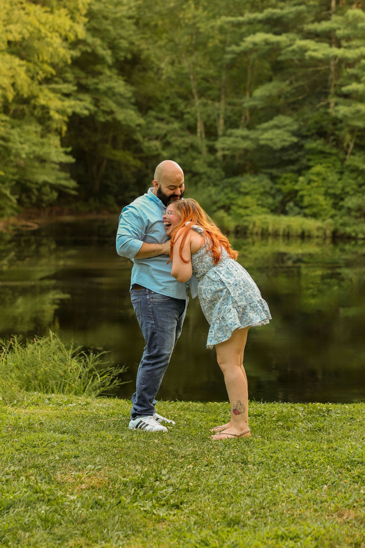 Couple sharing a tender moment by a reflective pond surrounded by vibrant green trees at golden hour.