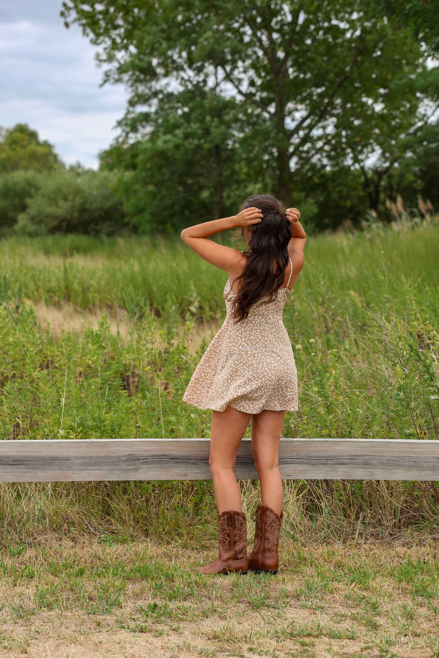 Young woman in floral mini dress and cowboy boots standing at wooden fence in meadow.