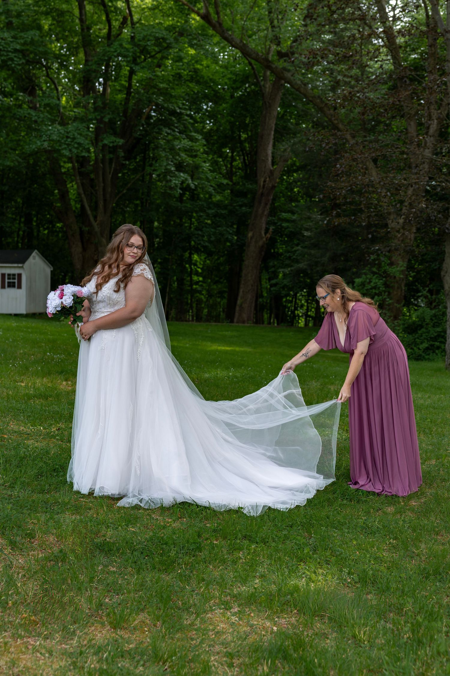 Bridesmaid in mauve dress arranges bride's white wedding veil and train on green grass lawn.
