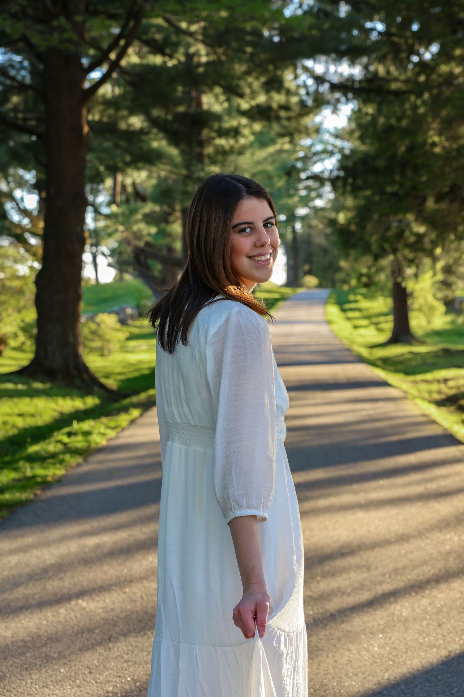 Young woman in white maxi dress smiling on tree-lined path in golden hour light.