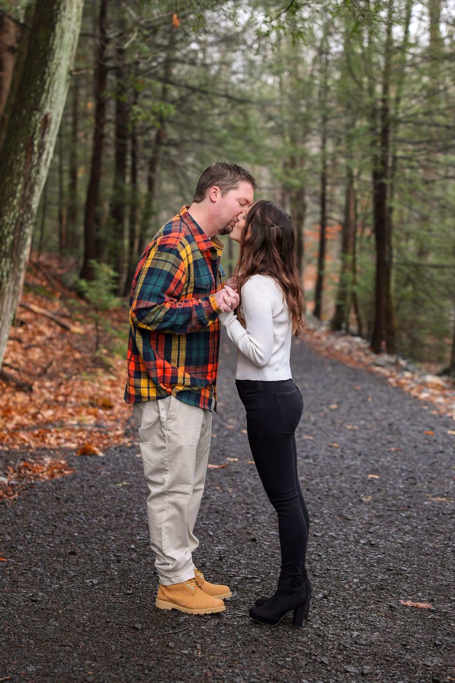 Romantic couple kissing while holding hands on a gravel forest path surrounded by tall trees in fall.