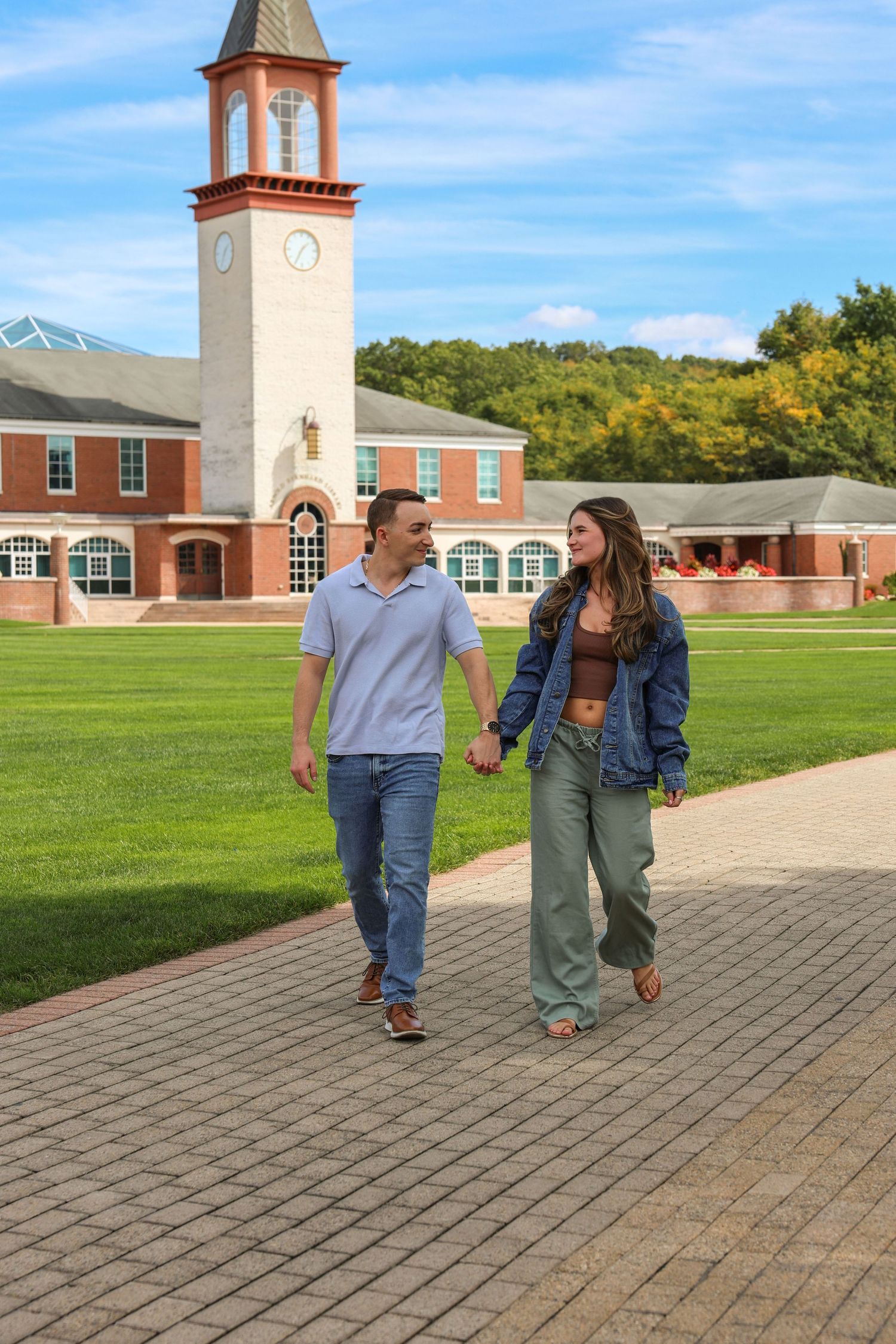 Engaged couple walking hand in hand on a brick campus pathway with a tall clock tower and green lawn.