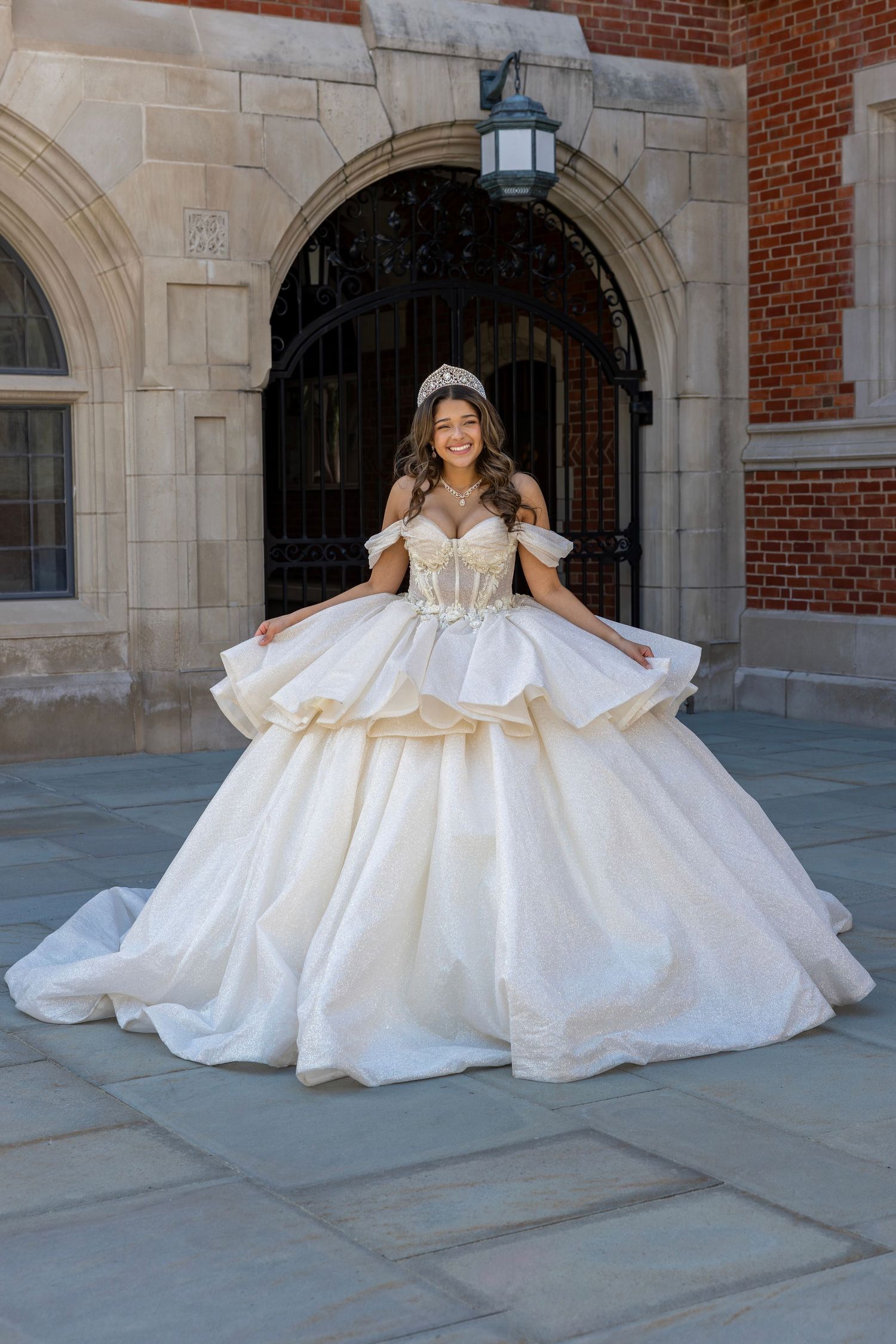 Joyful quinceañera in white and gold layered ball gown with tiara at stone archway.
