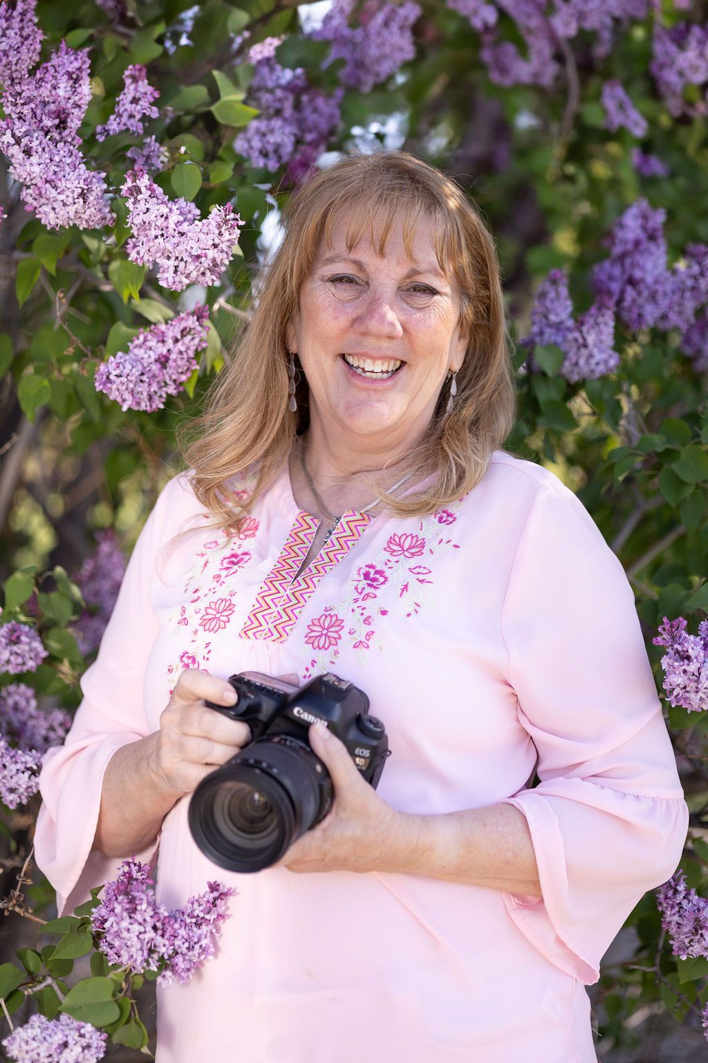 Lisa Craig - Lisa Craig Photography wearing pink embroidered top poses with her DSLR camera against blooming purple lilac bushes in spring.