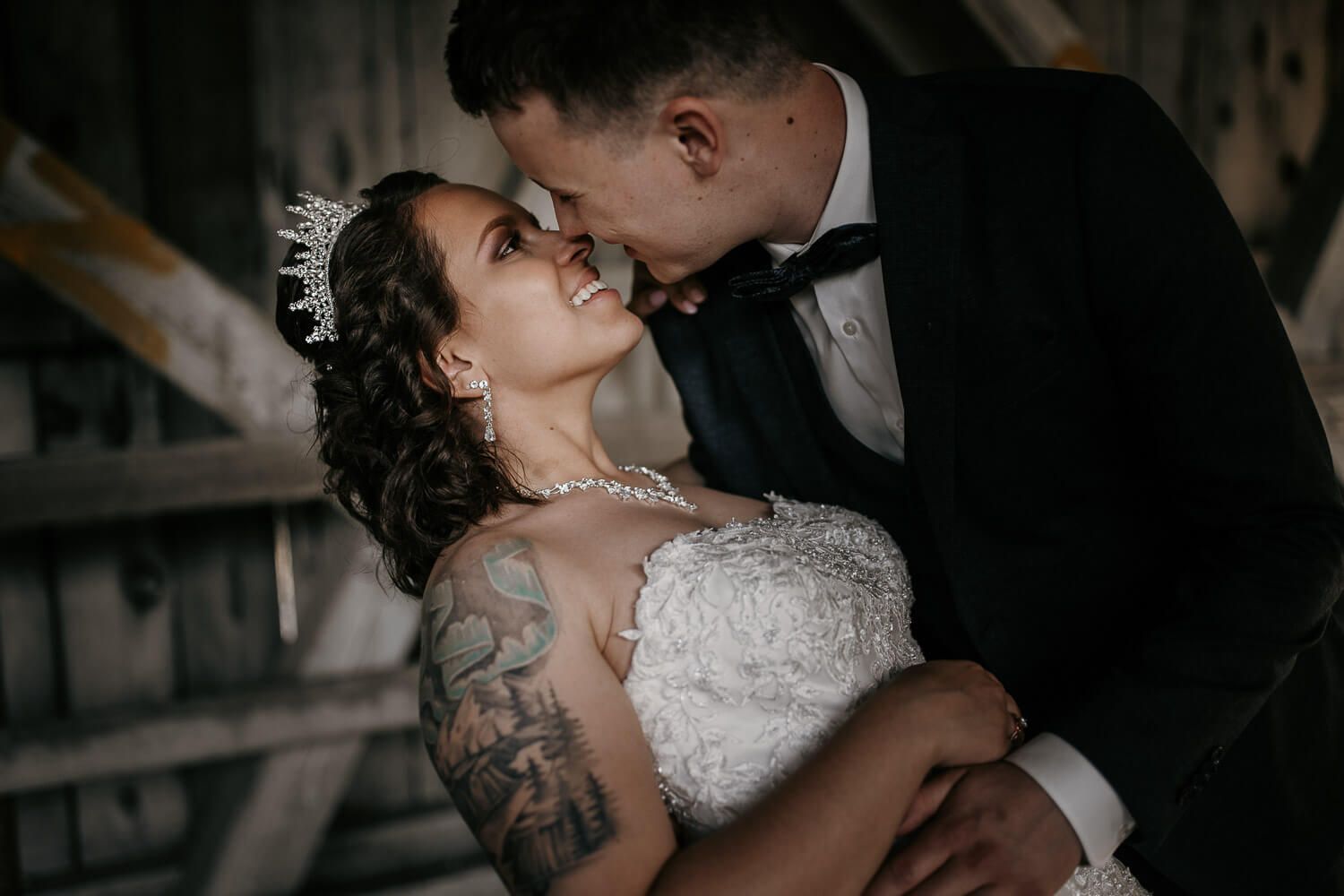 Bride and groom share a romantic moment, with the bride wearing a lace gown and tiara in a rustic setting.