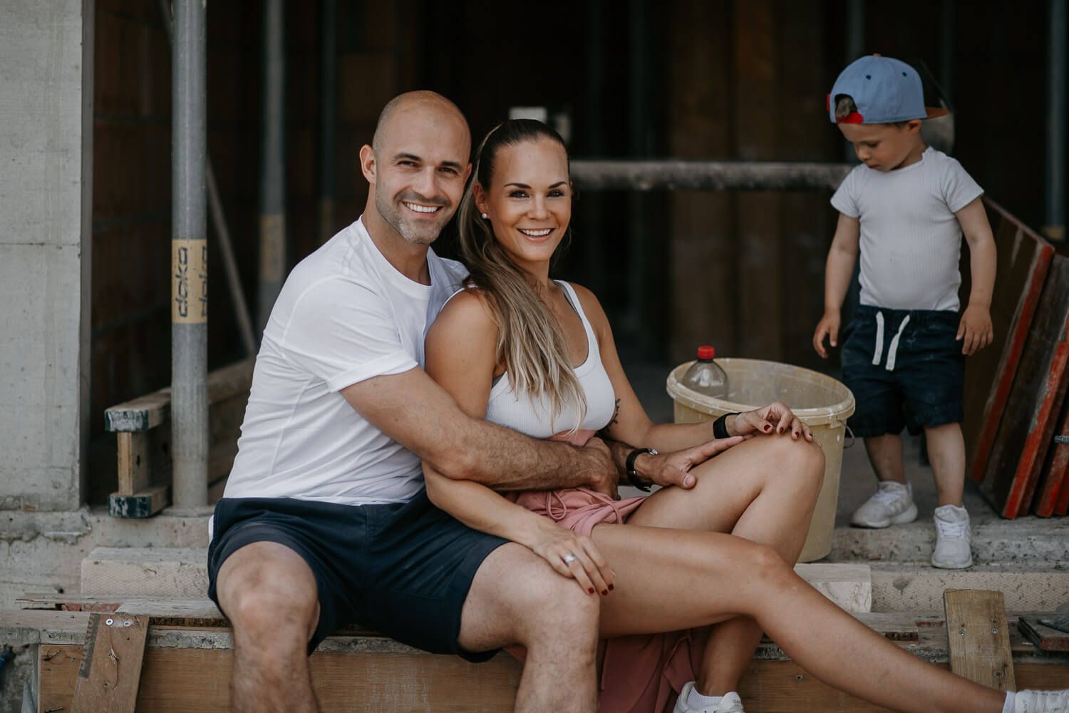 Smiling couple sitting together at construction site of new home with young child playing nearby.