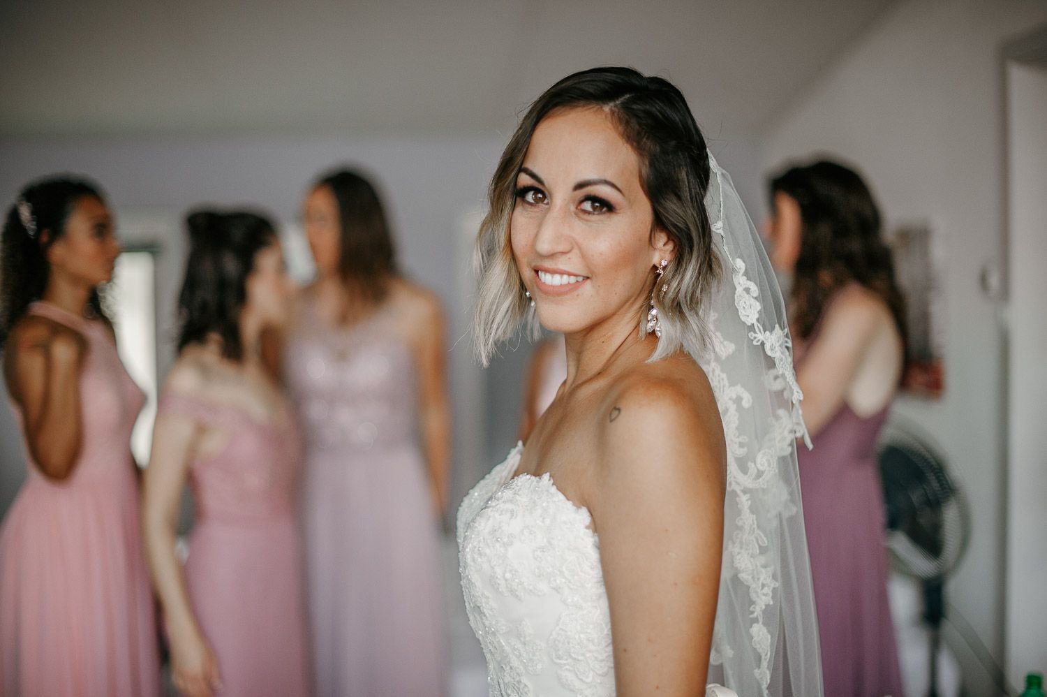 Smiling bride in white wedding dress and veil with bridesmaids in pink dresses in the background.