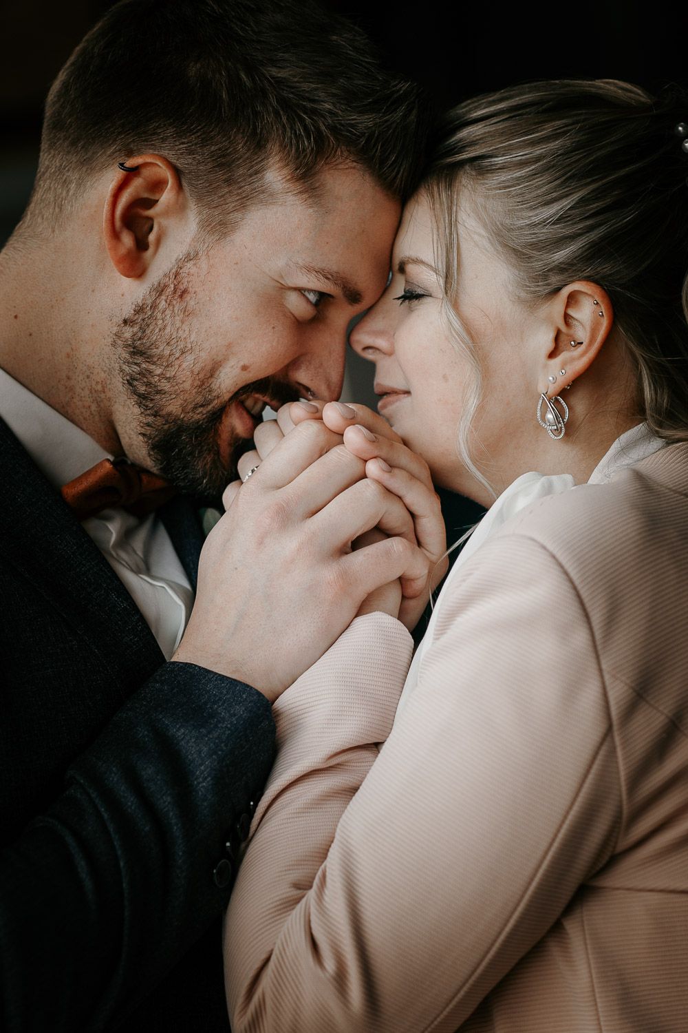 Intimate close-up of bearded groom and bride touching foreheads, holding hands tenderly in soft moody lighting.