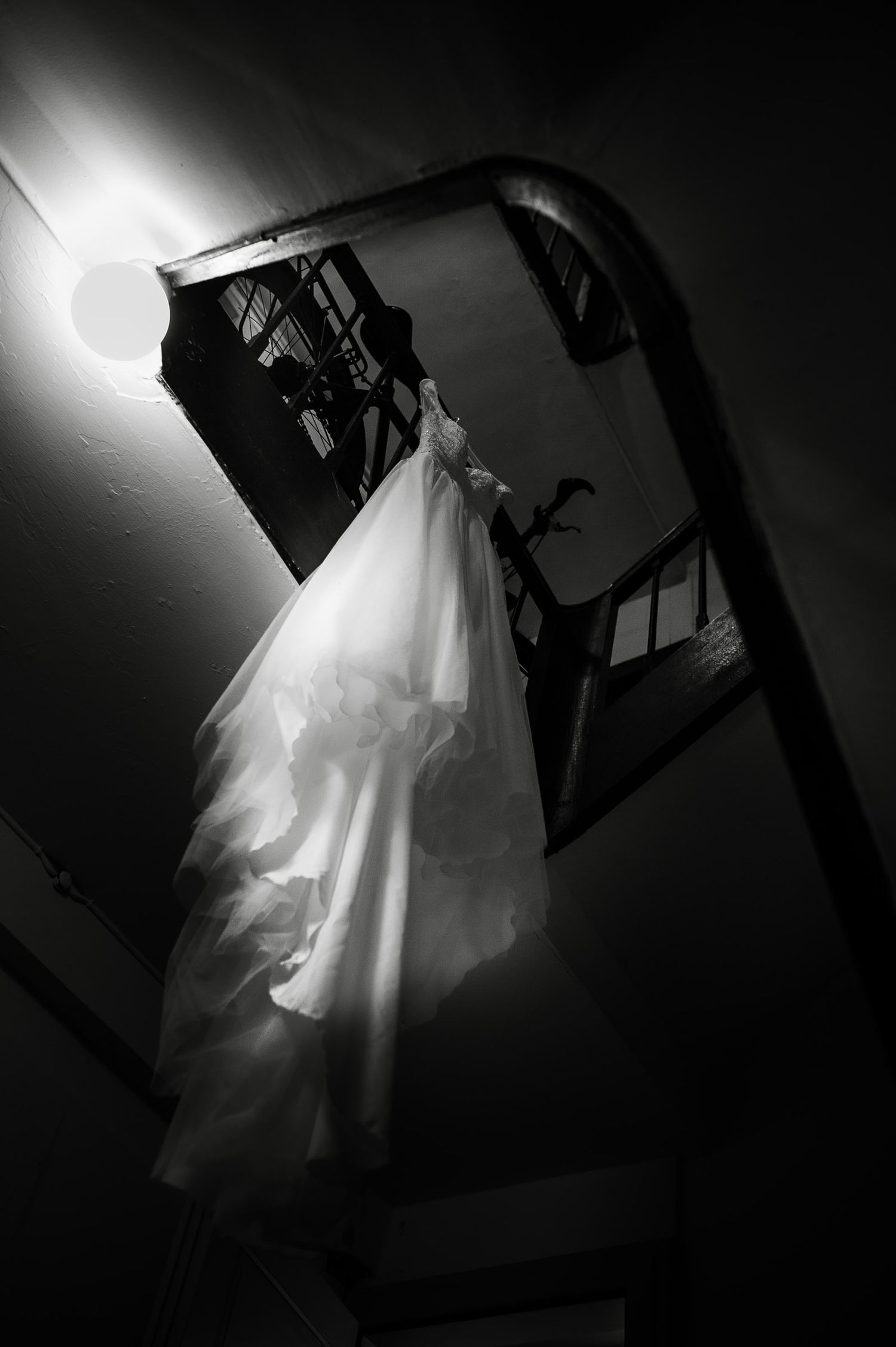 Black and white dramatic low-angle shot of a wedding dress hanging in a stairwell with light.