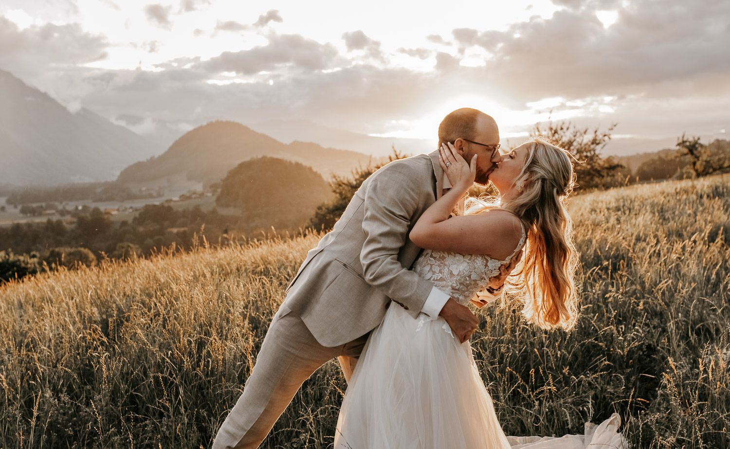 Romantic wedding couple kissing in golden field with mountain sunset backdrop.