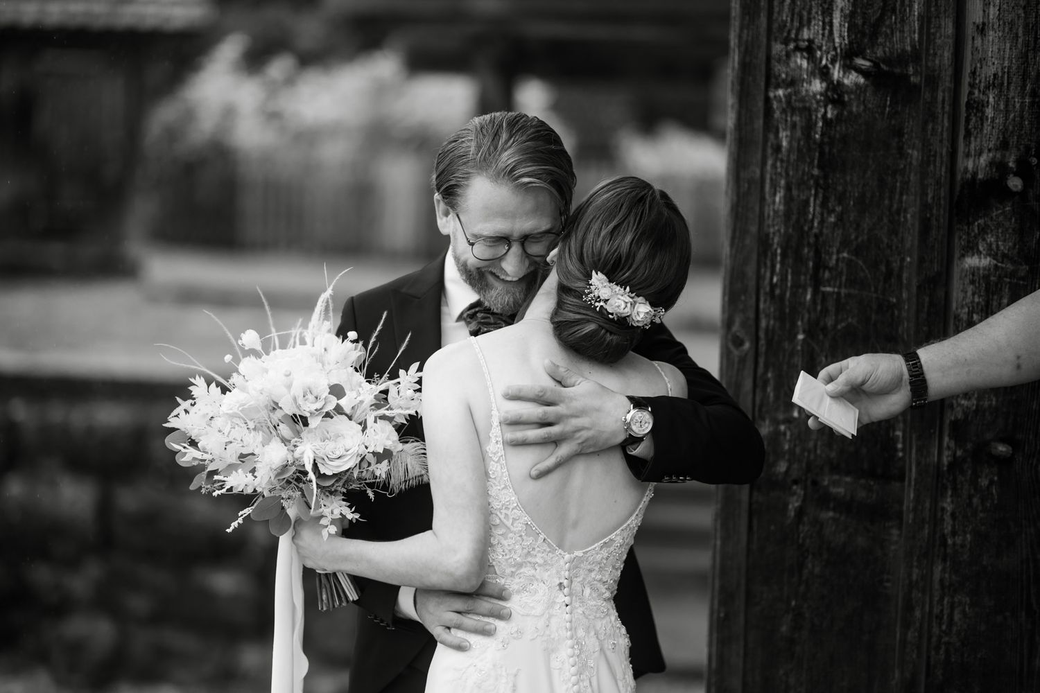 Black and white photo of groom embracing bride holding a floral bouquet at wedding ceremony.