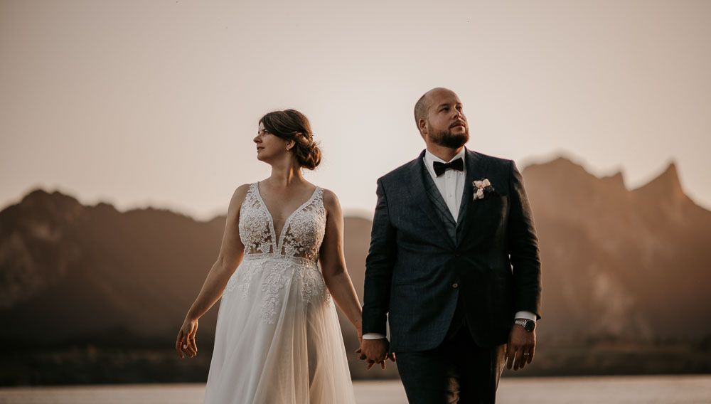 Elegant wedding couple holding hands walking at sunset with dramatic mountain and lake backdrop behind them.